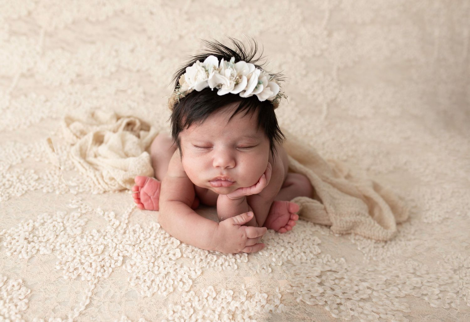 A sleeping newborn baby wearing a delicate white floral headband rests peacefully on cream-colored lace fabric.