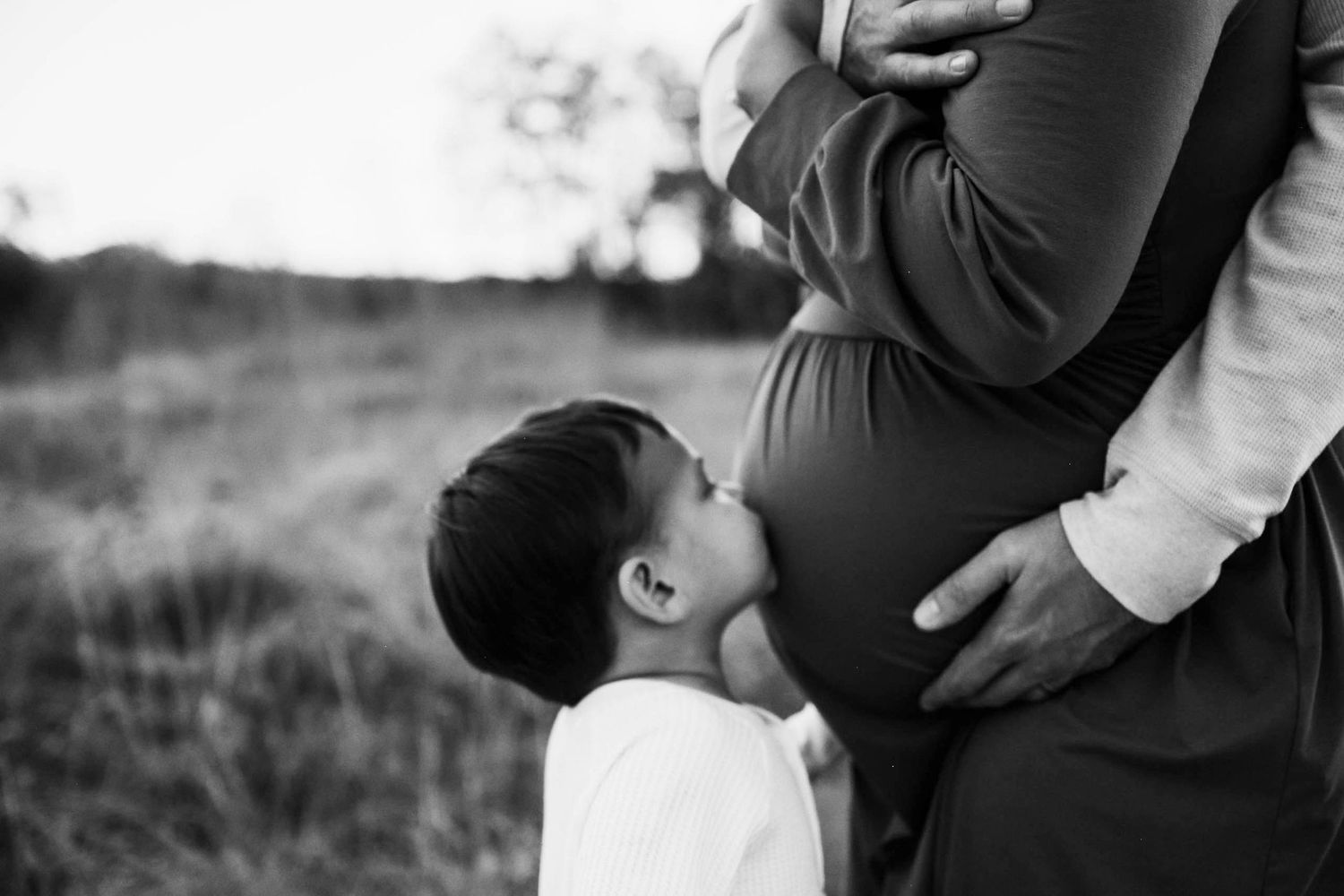 Black and white photo of a young child kissing a pregnant woman's belly in a field at sunset.