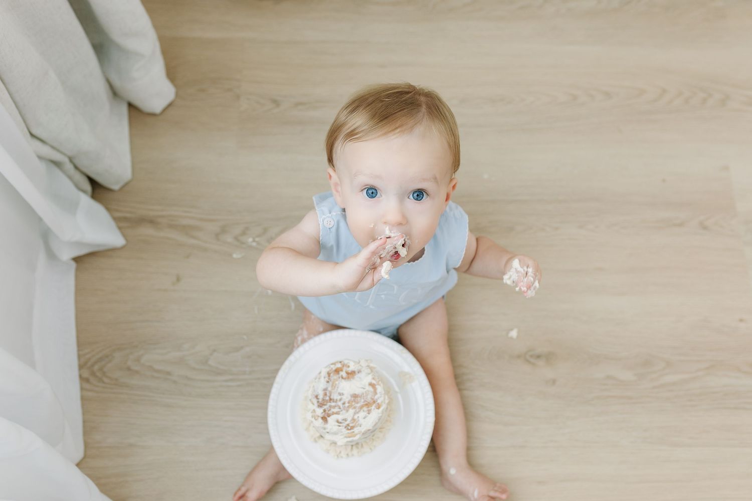 A blue-eyed baby in a light blue onesie sits on a wood floor, eating cake from a white plate.