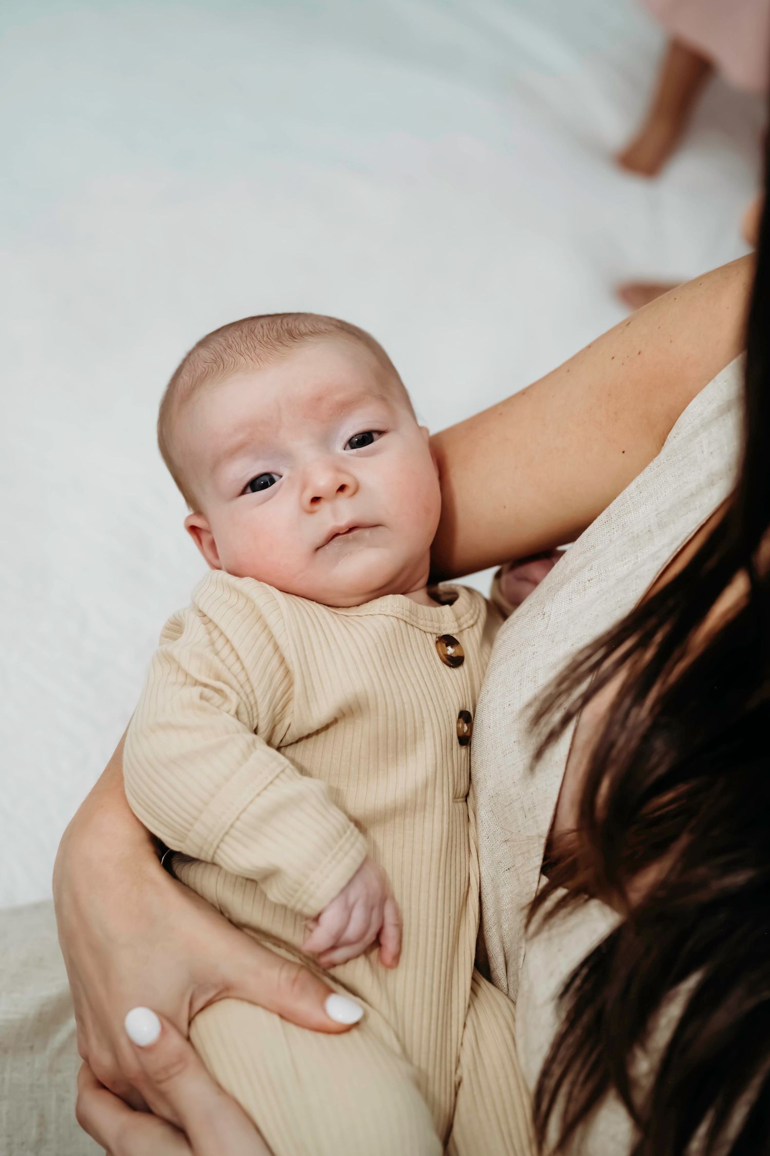 A baby wearing a cream colored outfit lies on white bedding during an intimate portrait session. Northlake tx, Lifestyle Newborn Session
