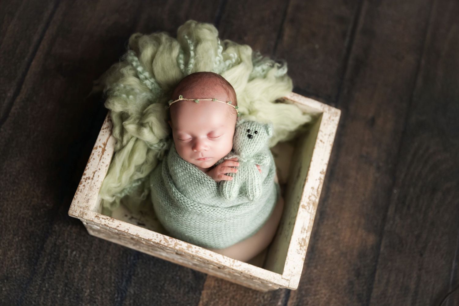 Newborn baby swaddled in green wrap sleeping in a wooden box with a tiny teddy bear for a professional photo session.