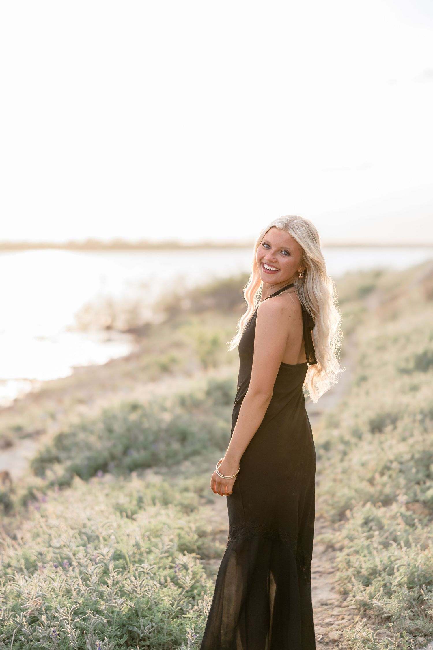 Smiling blonde woman in black maxi dress standing on a coastal hillside at golden hour.