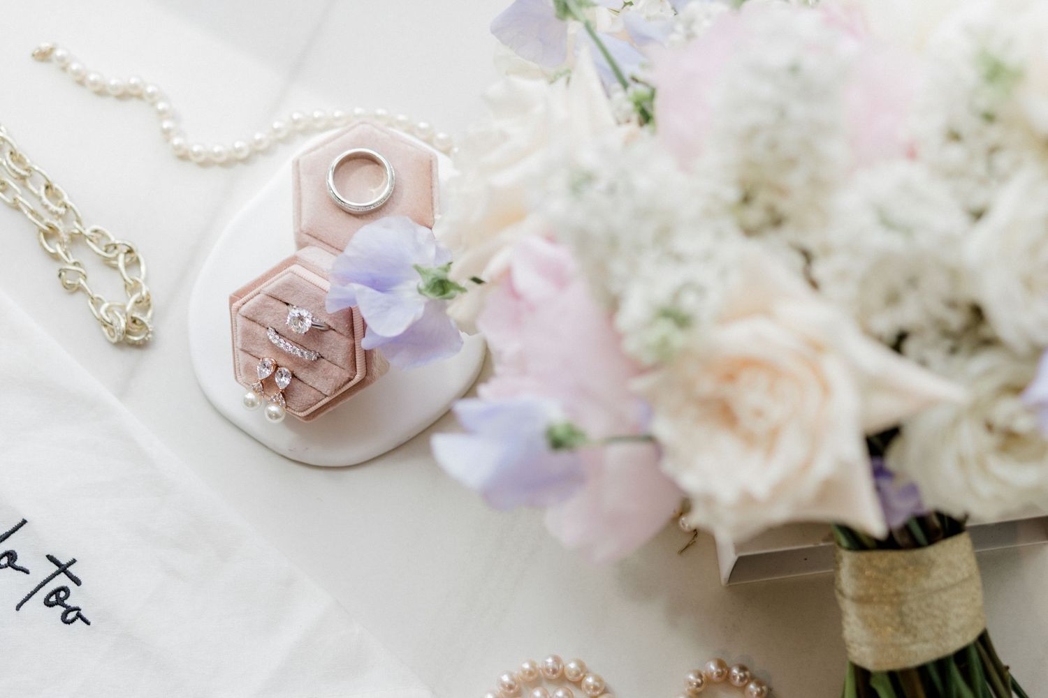 Elegant wedding flat lay with pink ring box, gold rings, pearl necklace, and a soft pastel floral bouquet.