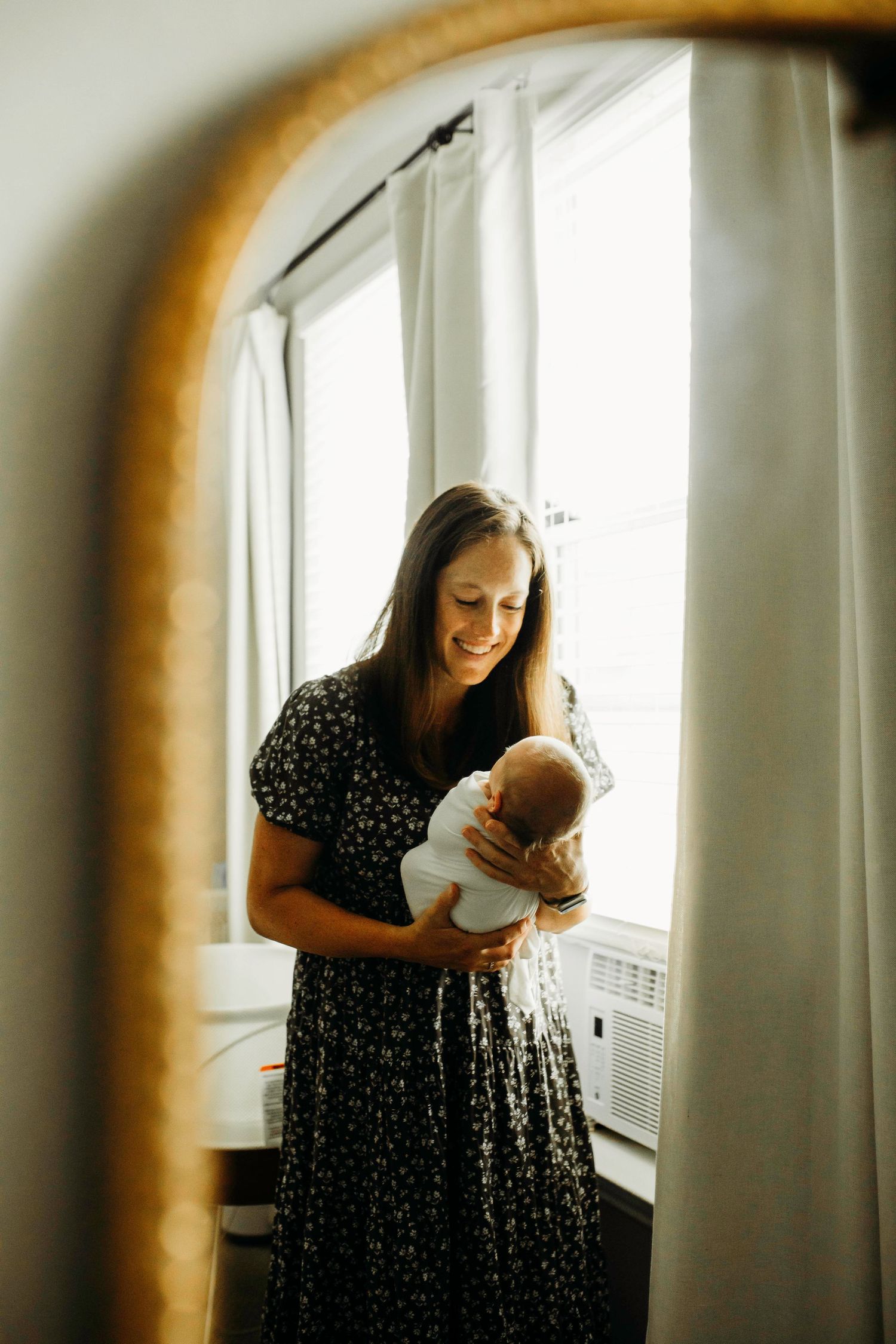 A tender moment captured through a mirror reflection shows a polka dot dress-clad figure holding a baby by a sunlit window.