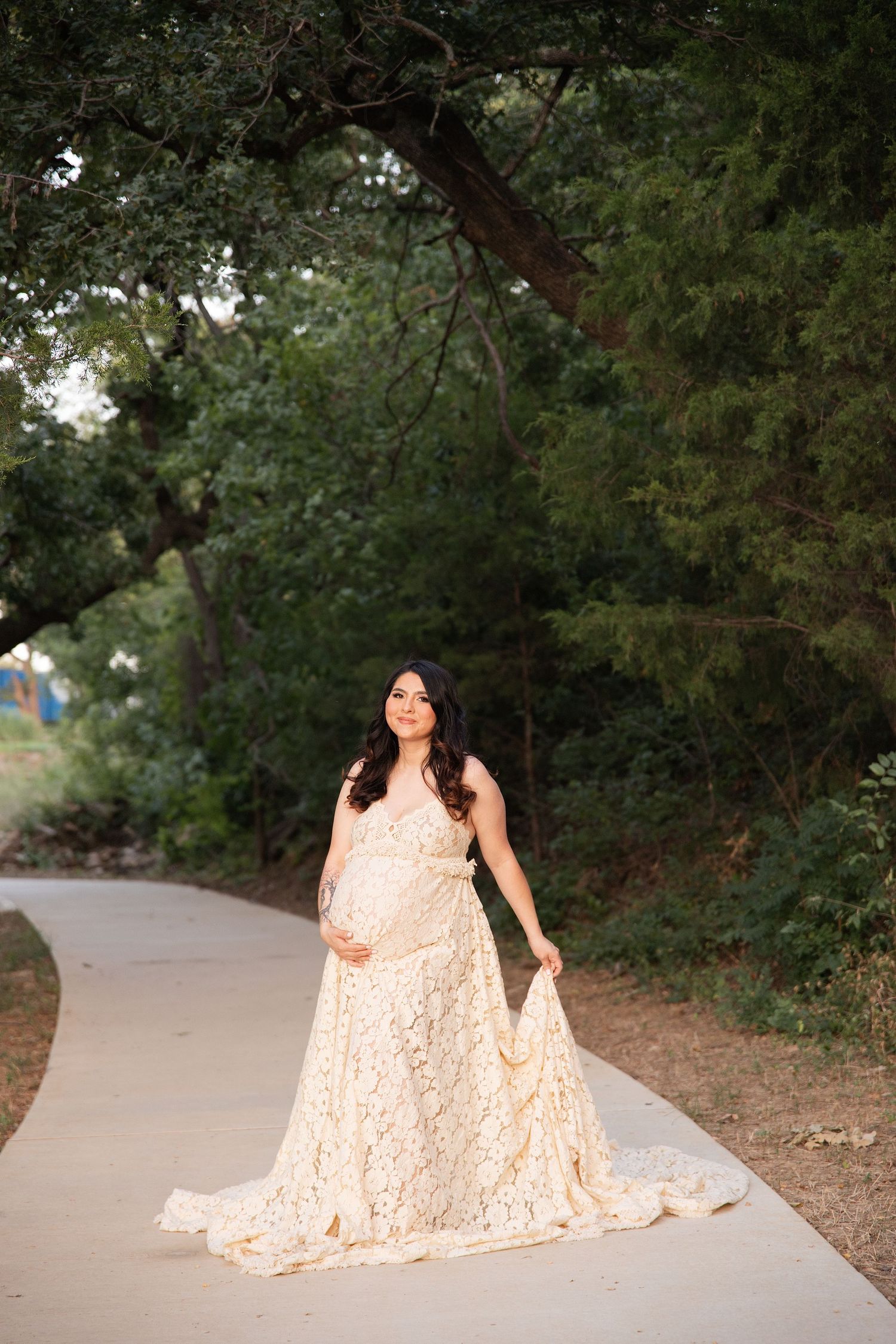 Pregnant woman in elegant cream lace maternity gown poses on a garden path surrounded by lush green trees.