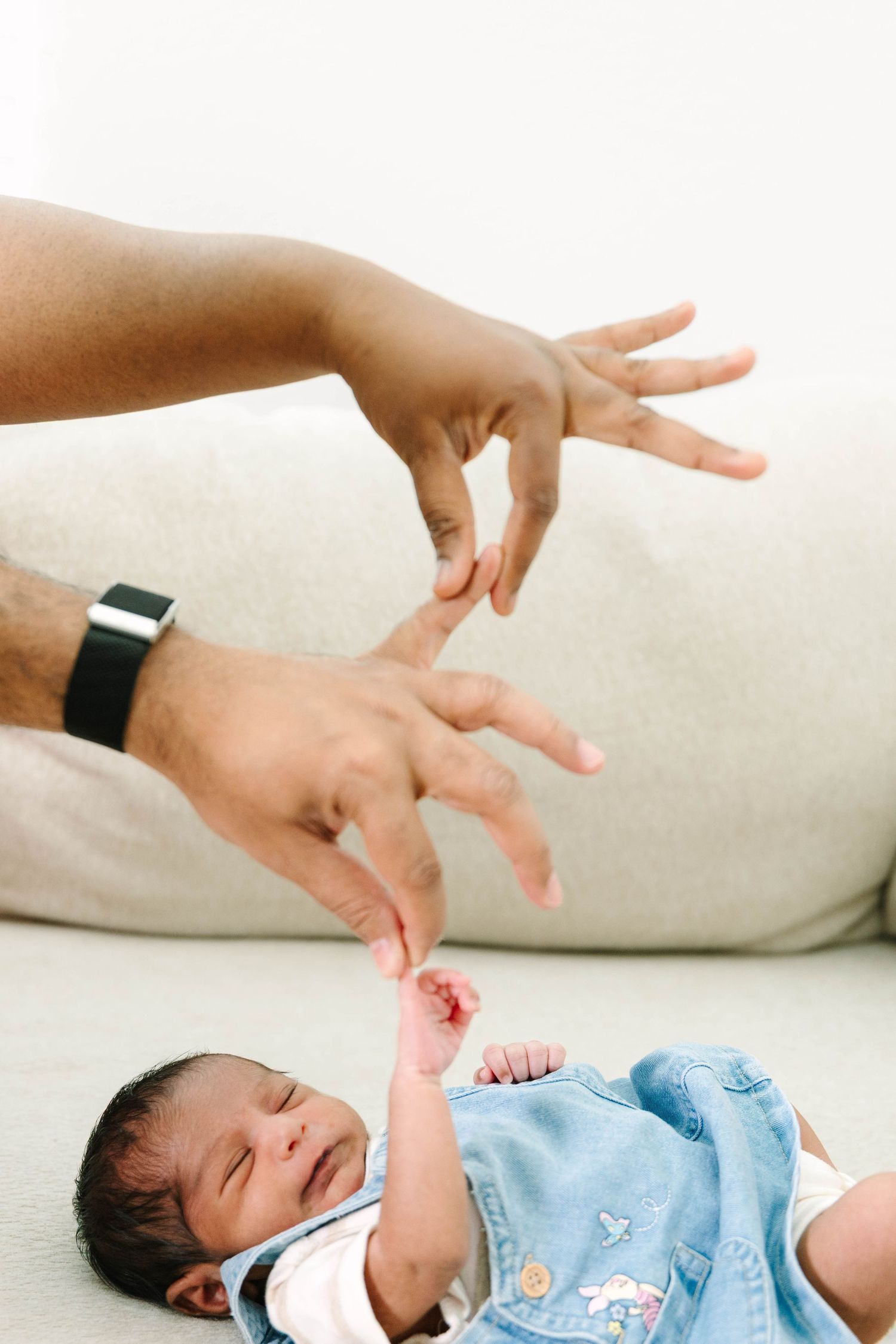 Parent's hands playfully reaching toward a sleeping newborn baby in a blue onesie on a soft surface.
