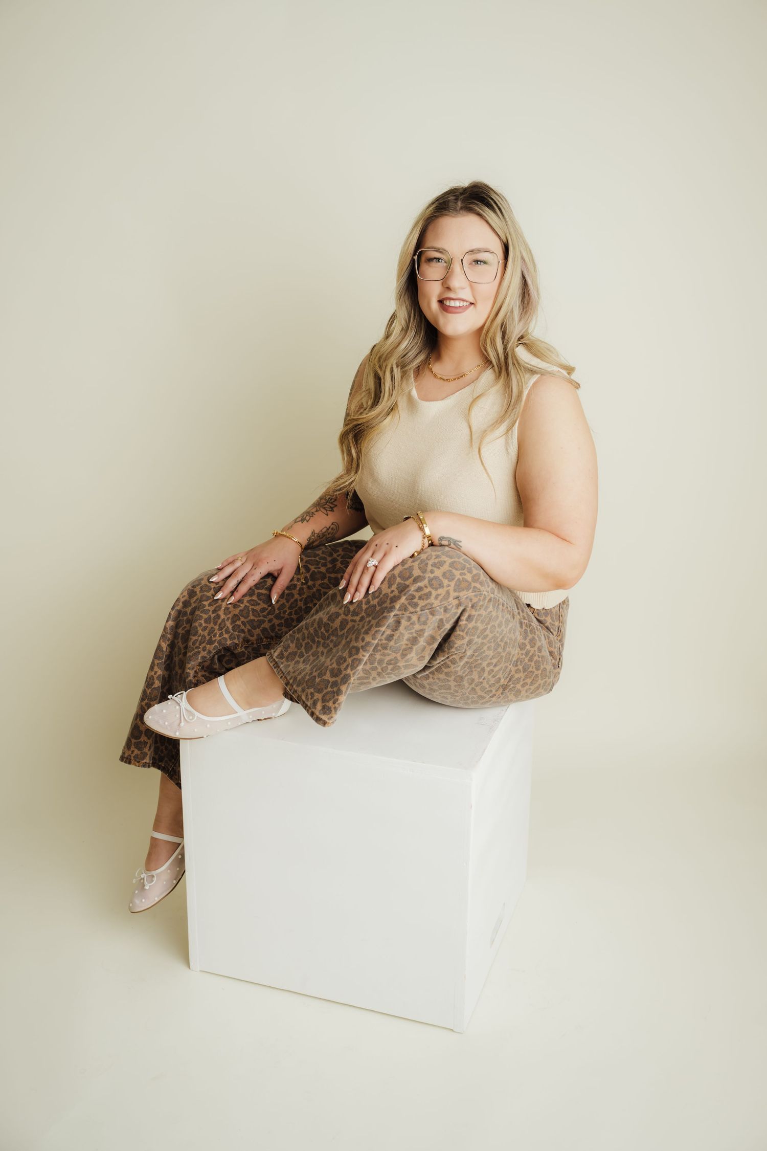 Smiling blonde woman in glasses, beige top, and leopard print pants sitting on a white cube in a studio.