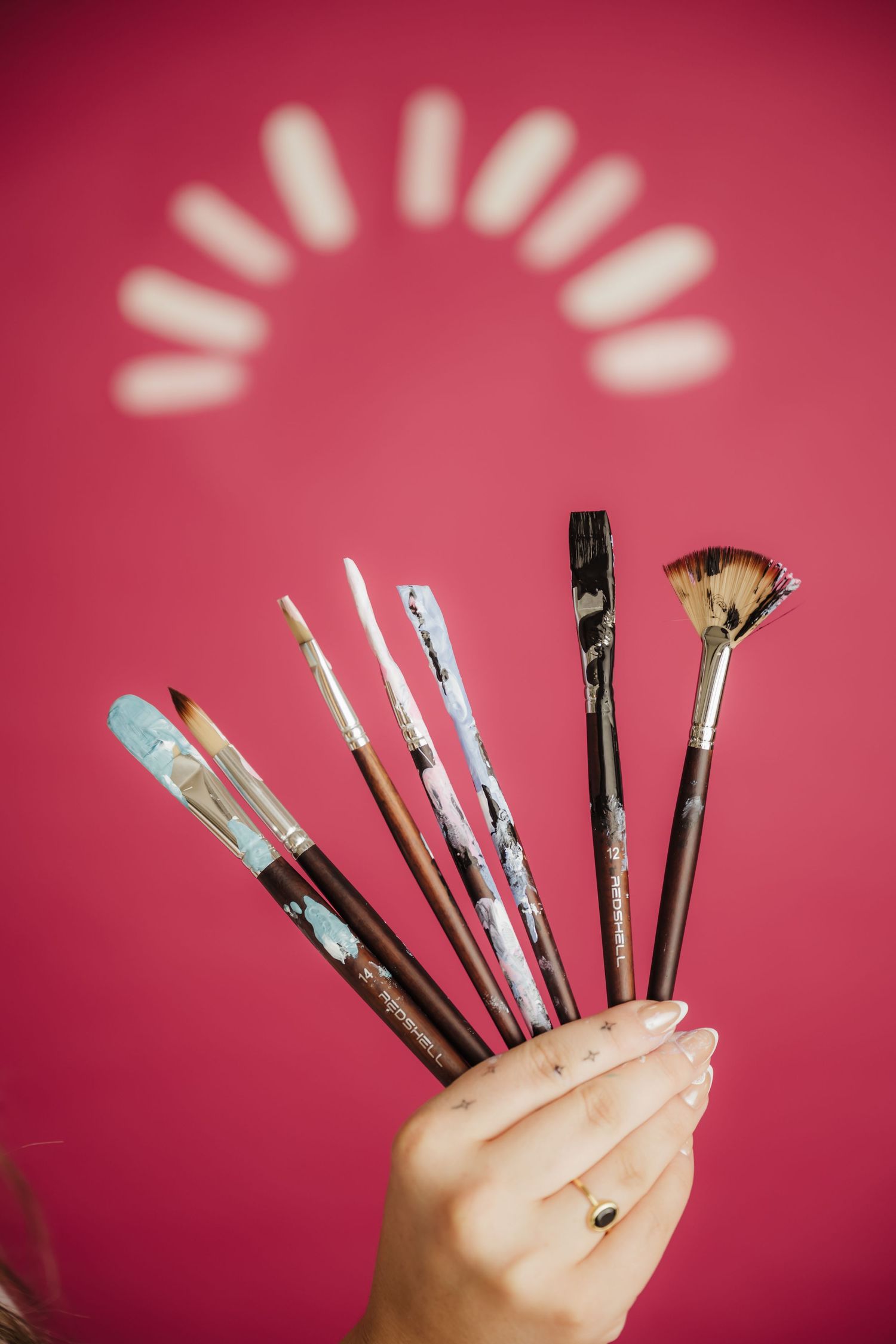 Hand holding assorted paint brushes with dried paint against a vibrant pink background with decorative light pattern.