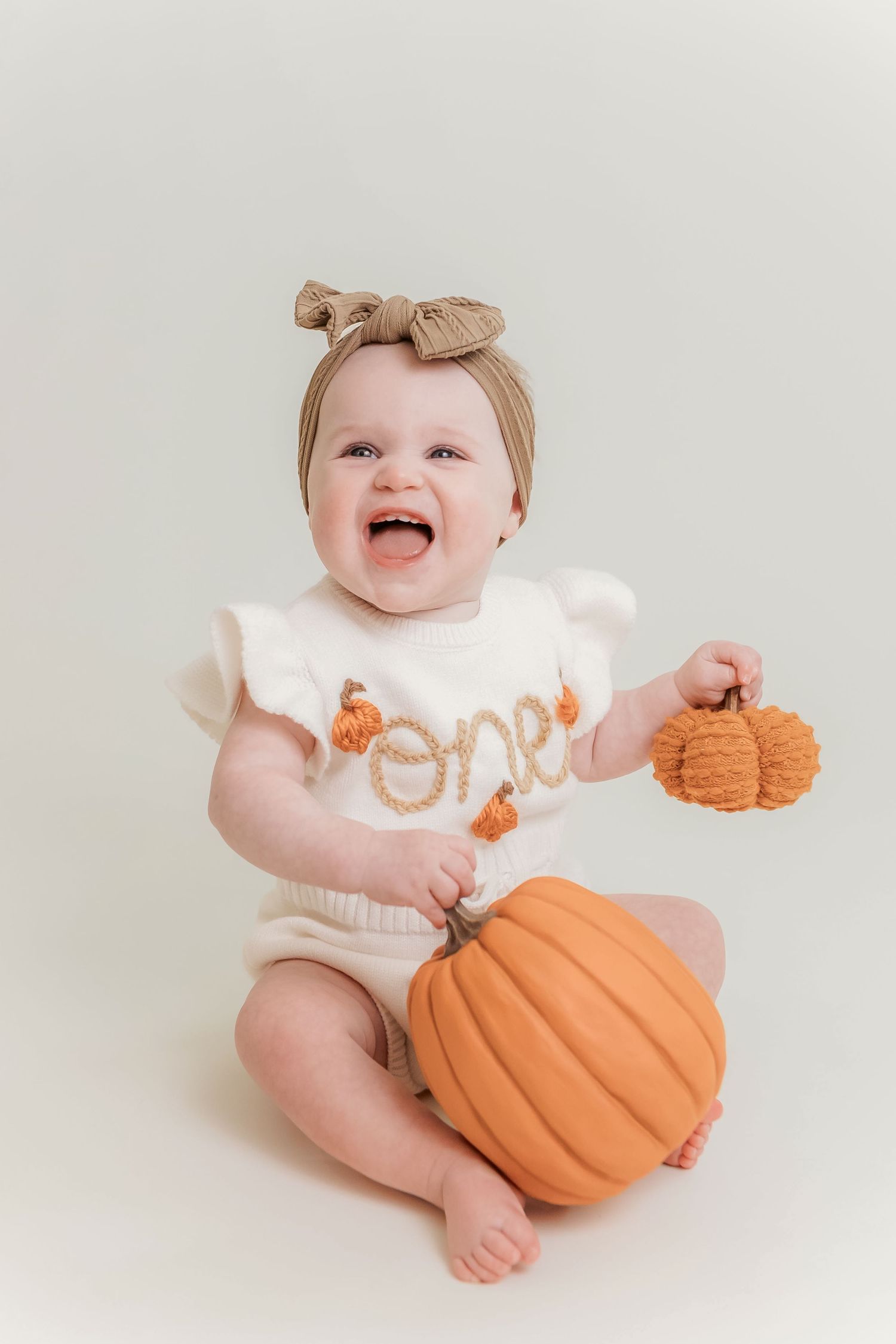 Baby girl in “one” pumpkin romper holding mini pumpkins, sitting beside orange pumpkin for first birthday photo.