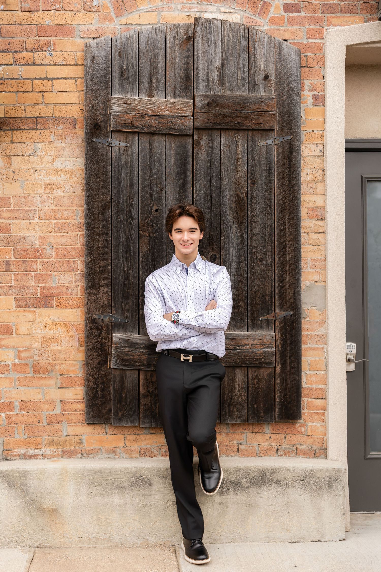 Young man in dress shirt and black pants stands confidently with arms crossed against a rustic brick wall with wooden doors.
