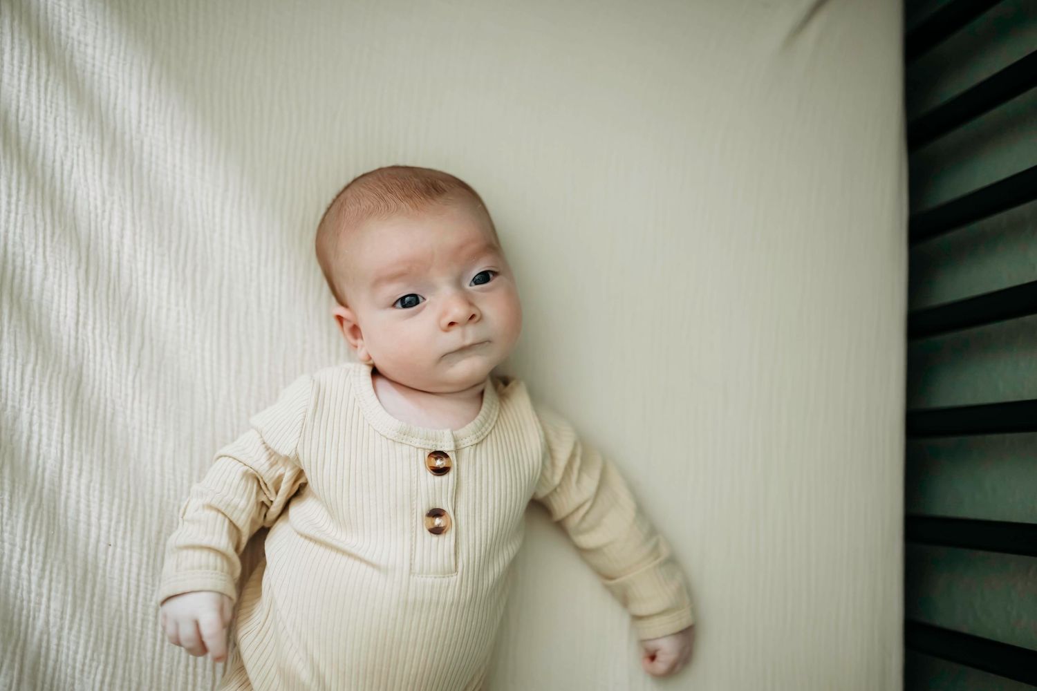 A baby in a cream colored ribbed outfit lies on light colored bedding looking up at the camera.