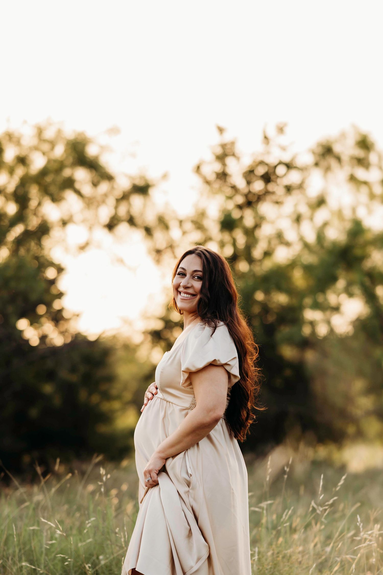 A mother and child embrace in a sunlit meadow at golden hour with tall grass and trees in the background.