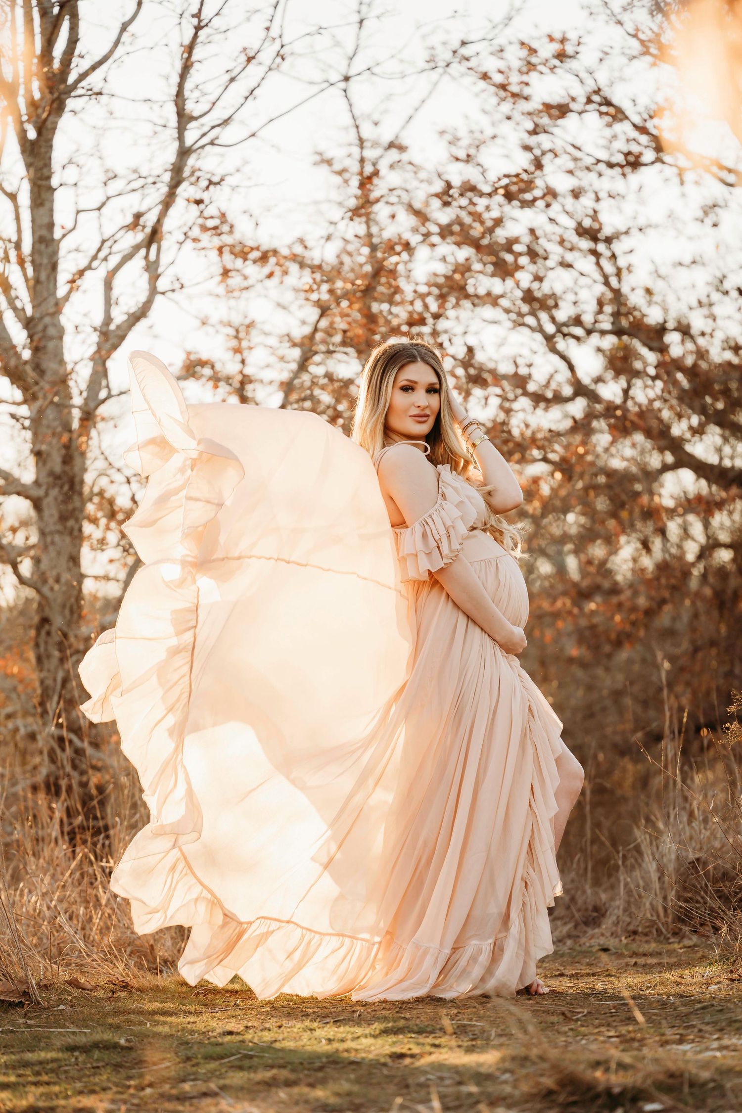 A flowing peach gown billows dramatically in golden sunlight against an autumn forest backdrop.
