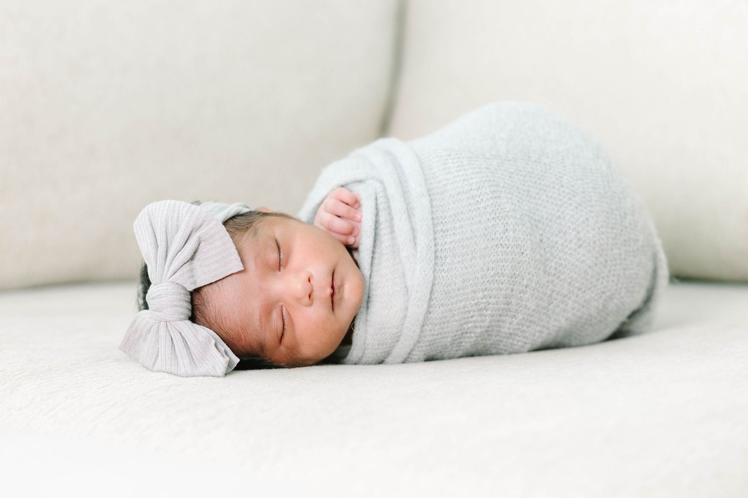 Sleeping newborn baby swaddled in a soft gray wrap with a bow headband on a white cushion.