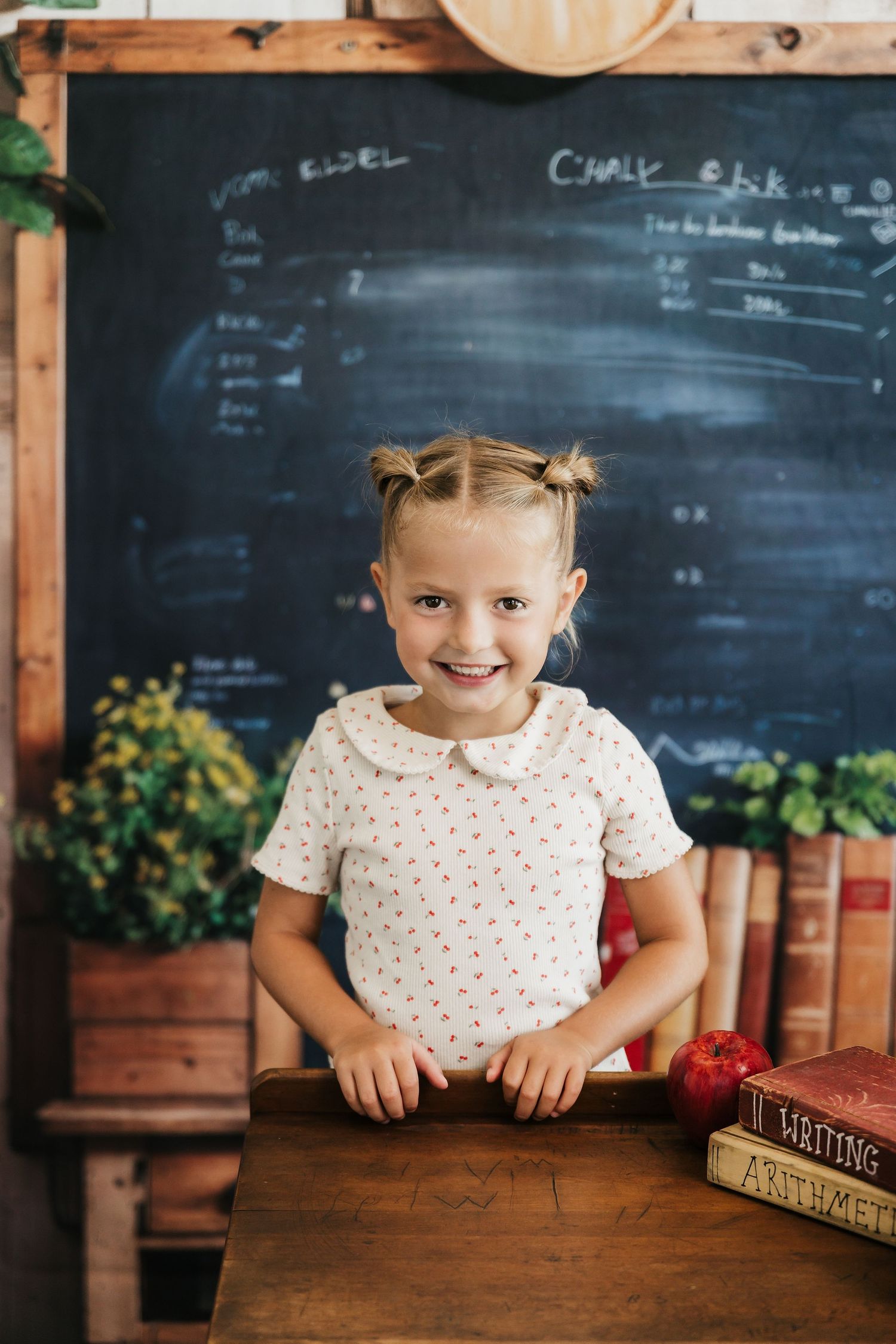 Smiling young girl with pigtails stands at a wooden desk with books and an apple in front of a chalkboard.