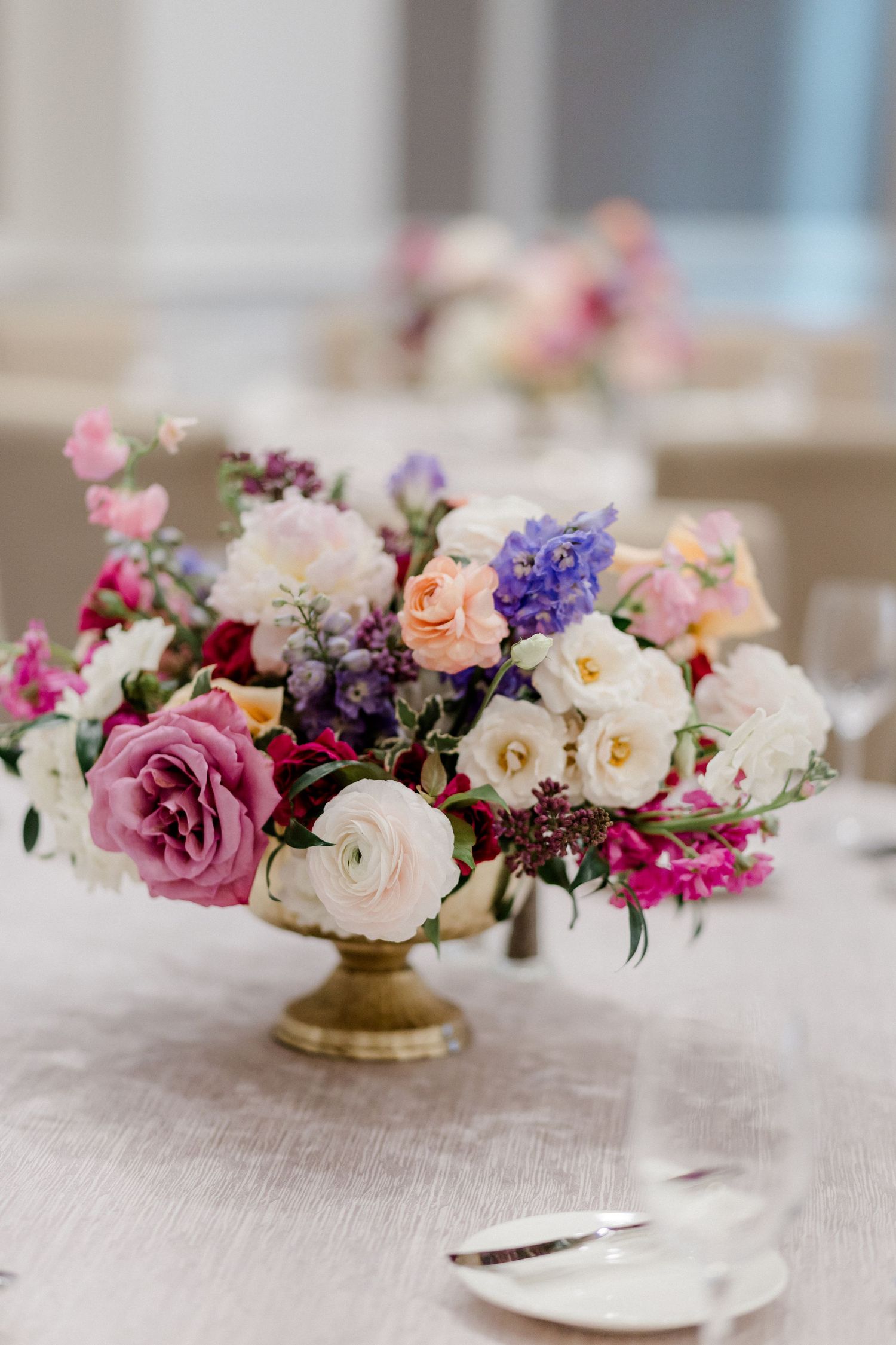 Gold pedestal vase with lush pink, white, and purple floral centerpiece on elegant table setting.