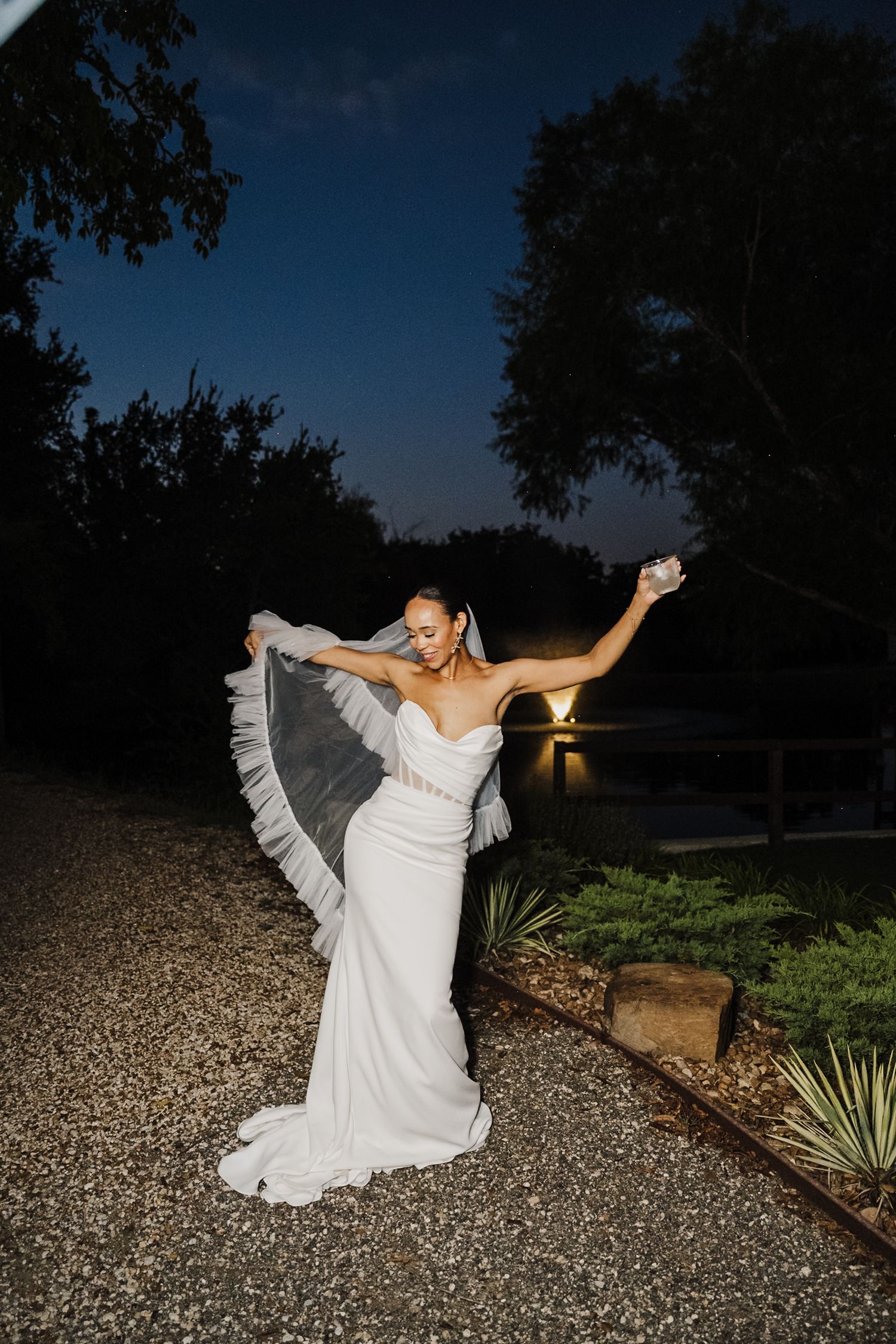 A bride in a white wedding dress twirls gracefully on a gravel path at dusk, creating a flowing motion effect.