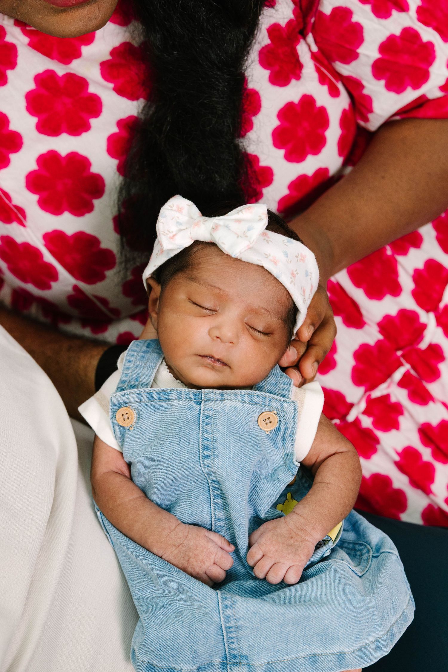 Sleeping newborn baby girl in denim dress and white bow headband, cradled by mother in red floral top.