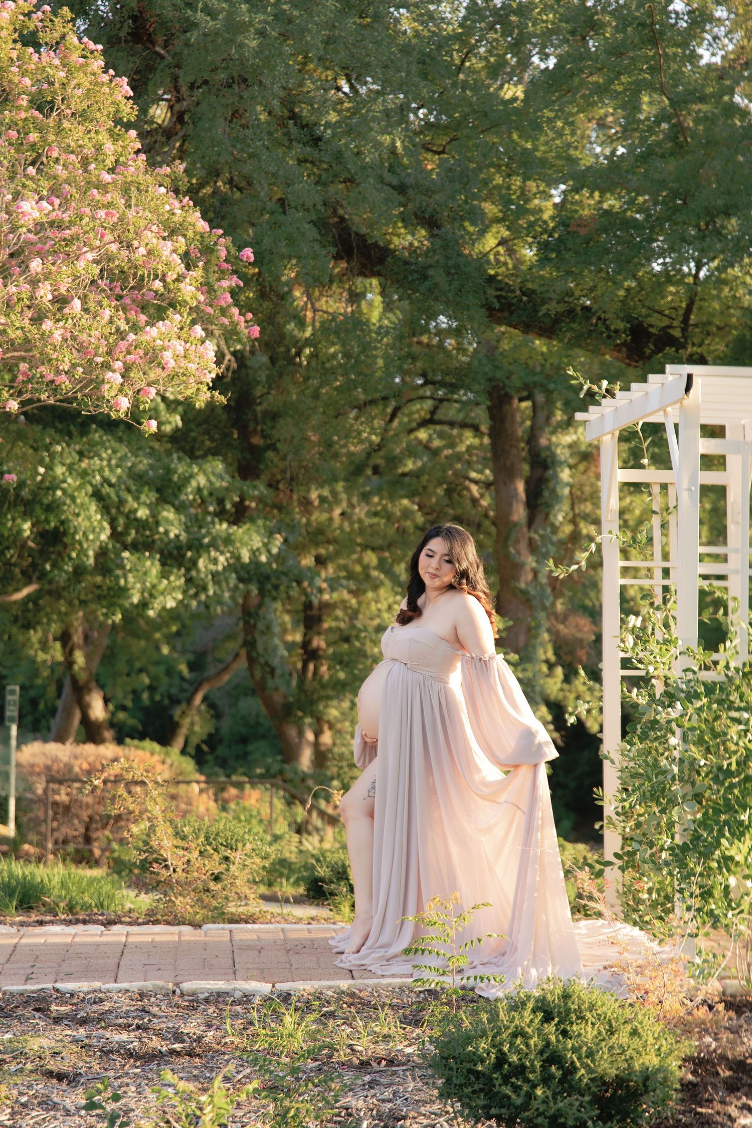 Pregnant woman in flowing blush gown poses gracefully in lush garden near white arbor and blooming trees.