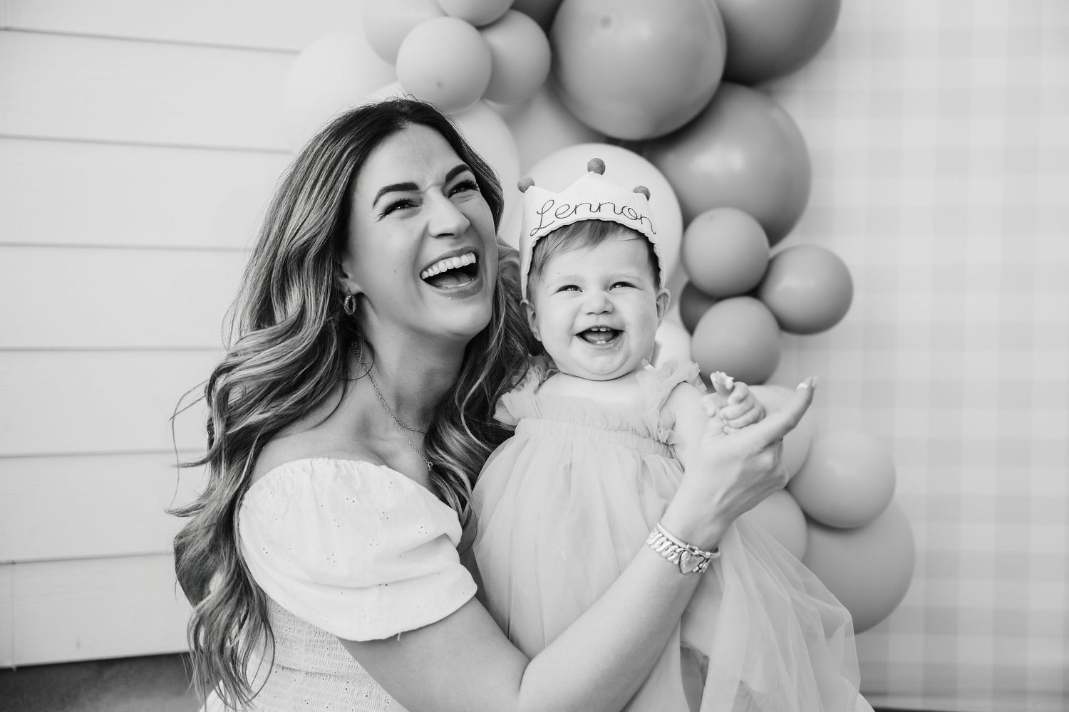 Black and white photo of a laughing woman holding a smiling baby girl at a balloon backdrop birthday party.
