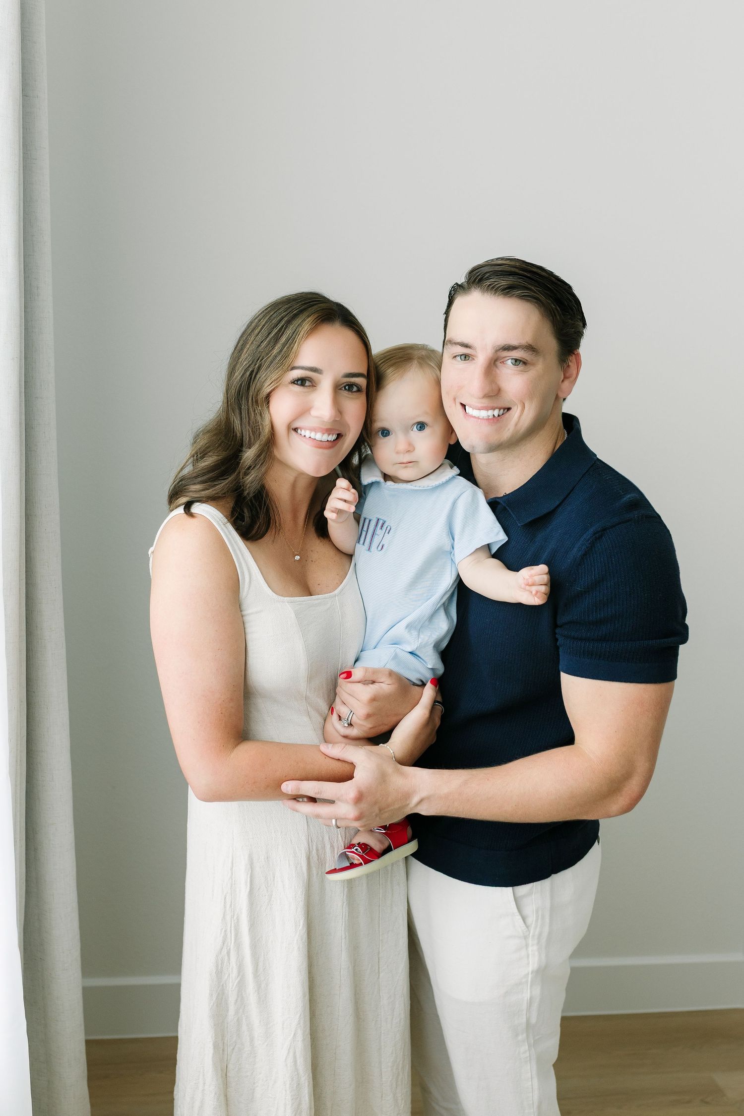 Smiling young family with mother in white dress and father in navy polo holding their baby in a bright studio.