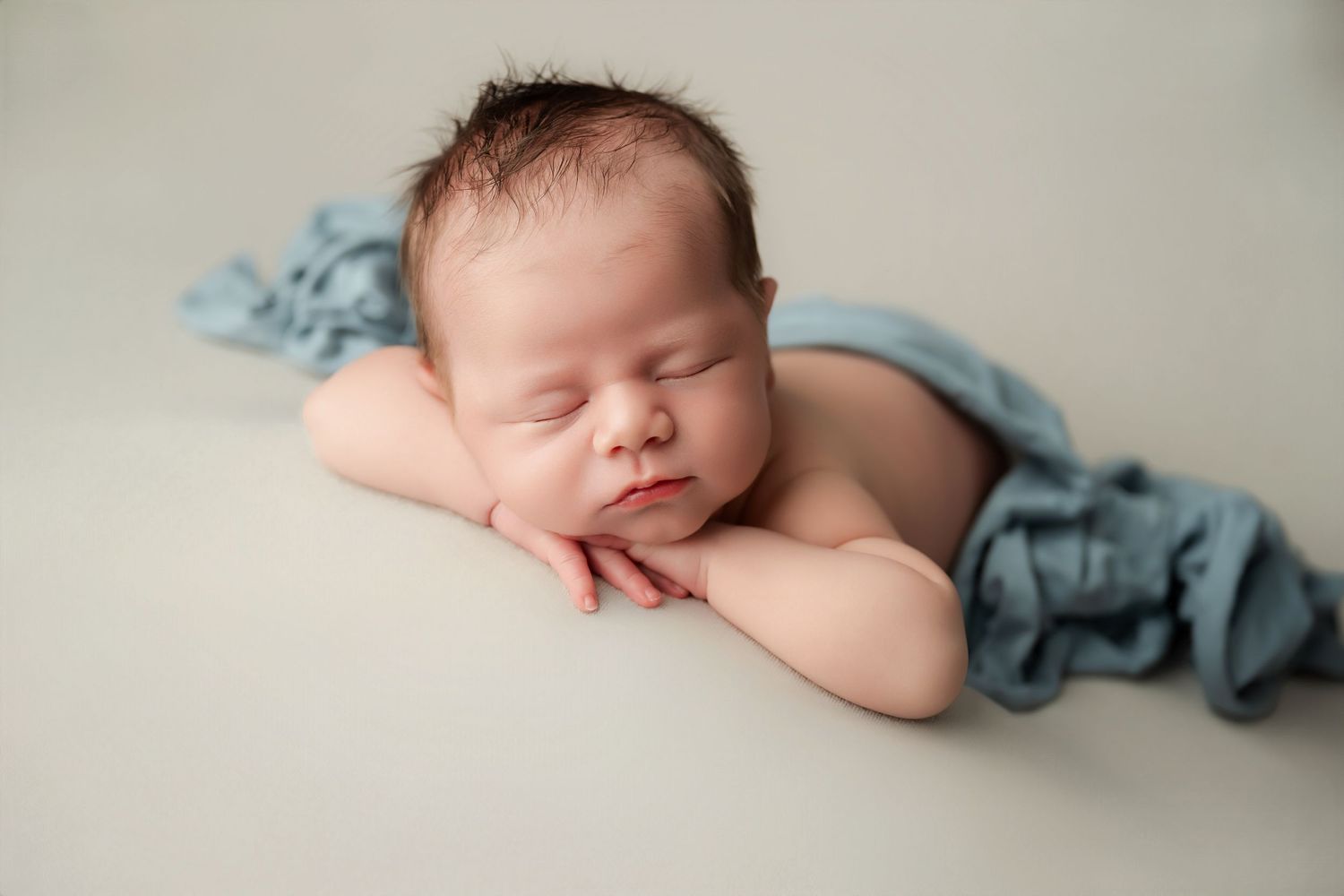 Peaceful newborn baby sleeping on white backdrop with blue fabric draped over, hands tucked under chin.