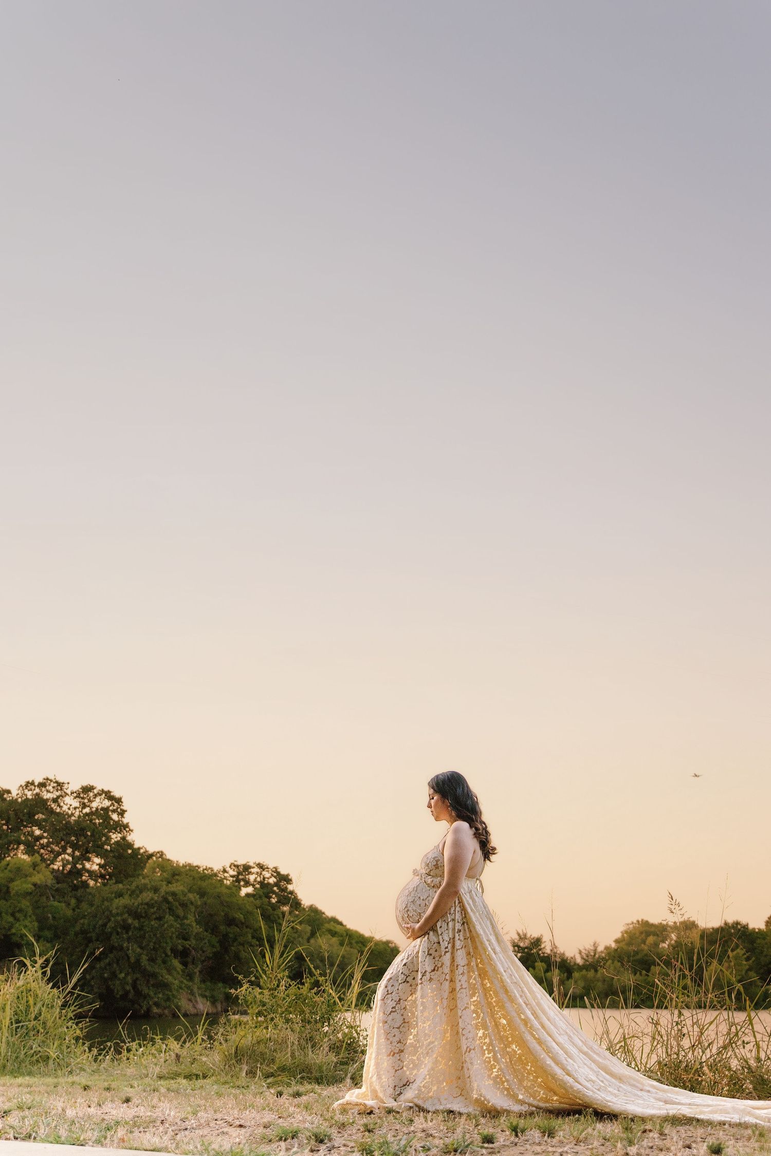 Pregnant woman in elegant lace gown cradles baby bump at golden hour near a serene lake surrounded by lush trees.