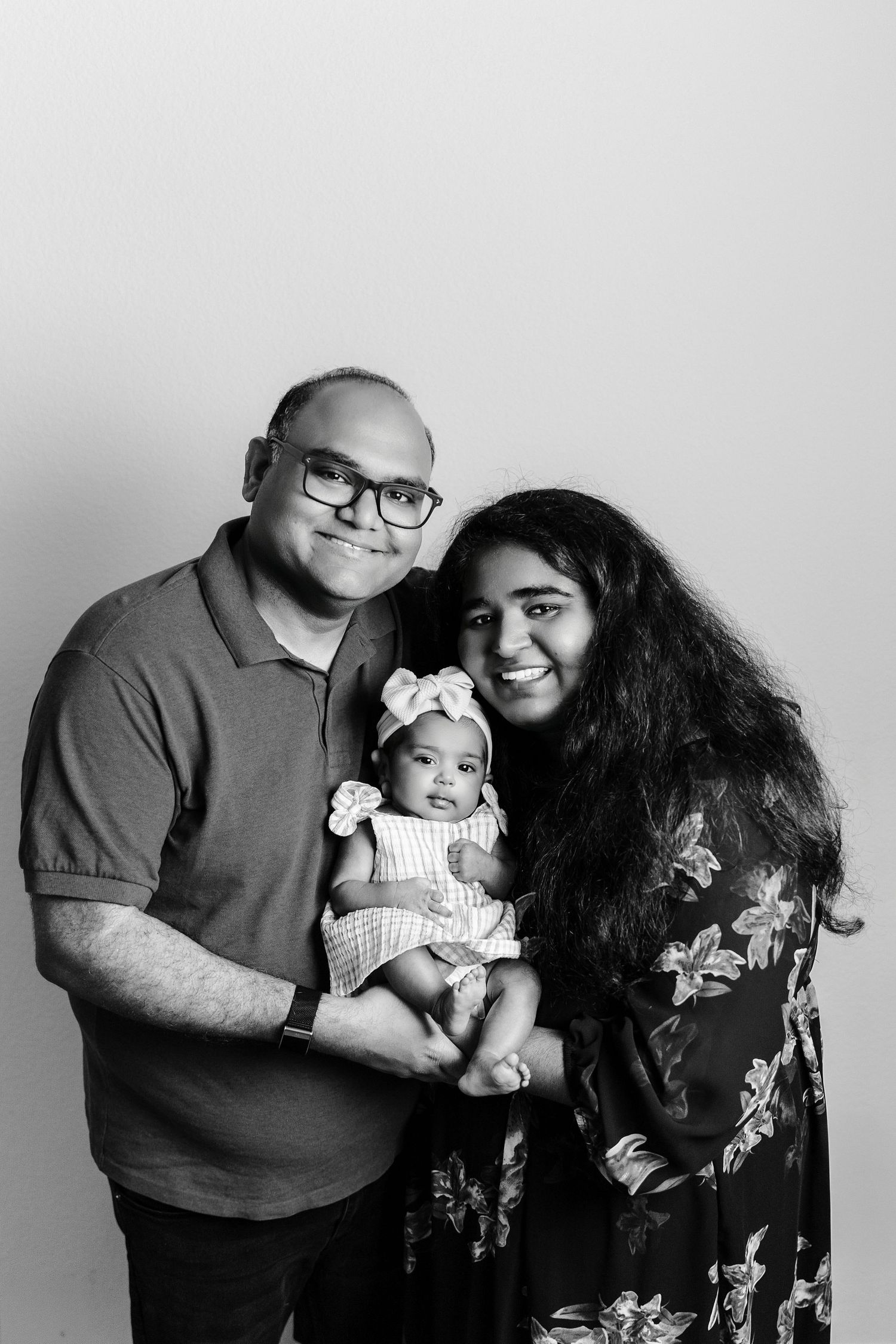 Black and white family portrait of parents smiling and holding their newborn baby dressed in a white outfit with a bow headband.