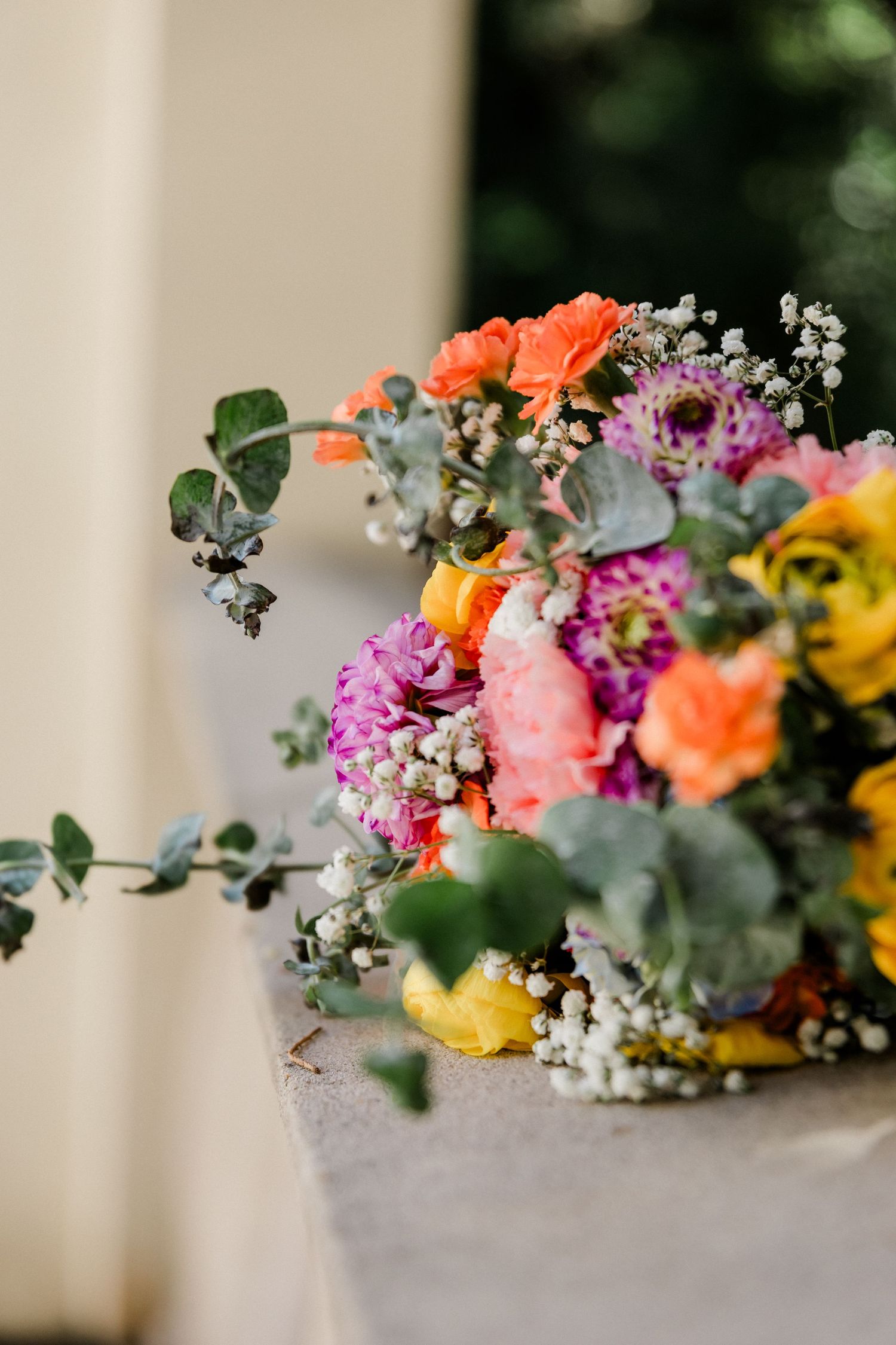 Vibrant wedding bouquet with orange, pink, and yellow flowers, eucalyptus, and baby's breath on a stone ledge.