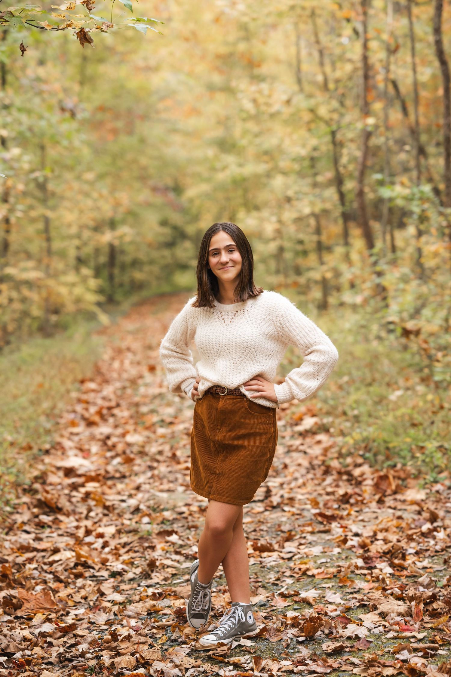 Person wearing cream sweater and brown skirt poses on leaf-covered forest trail.