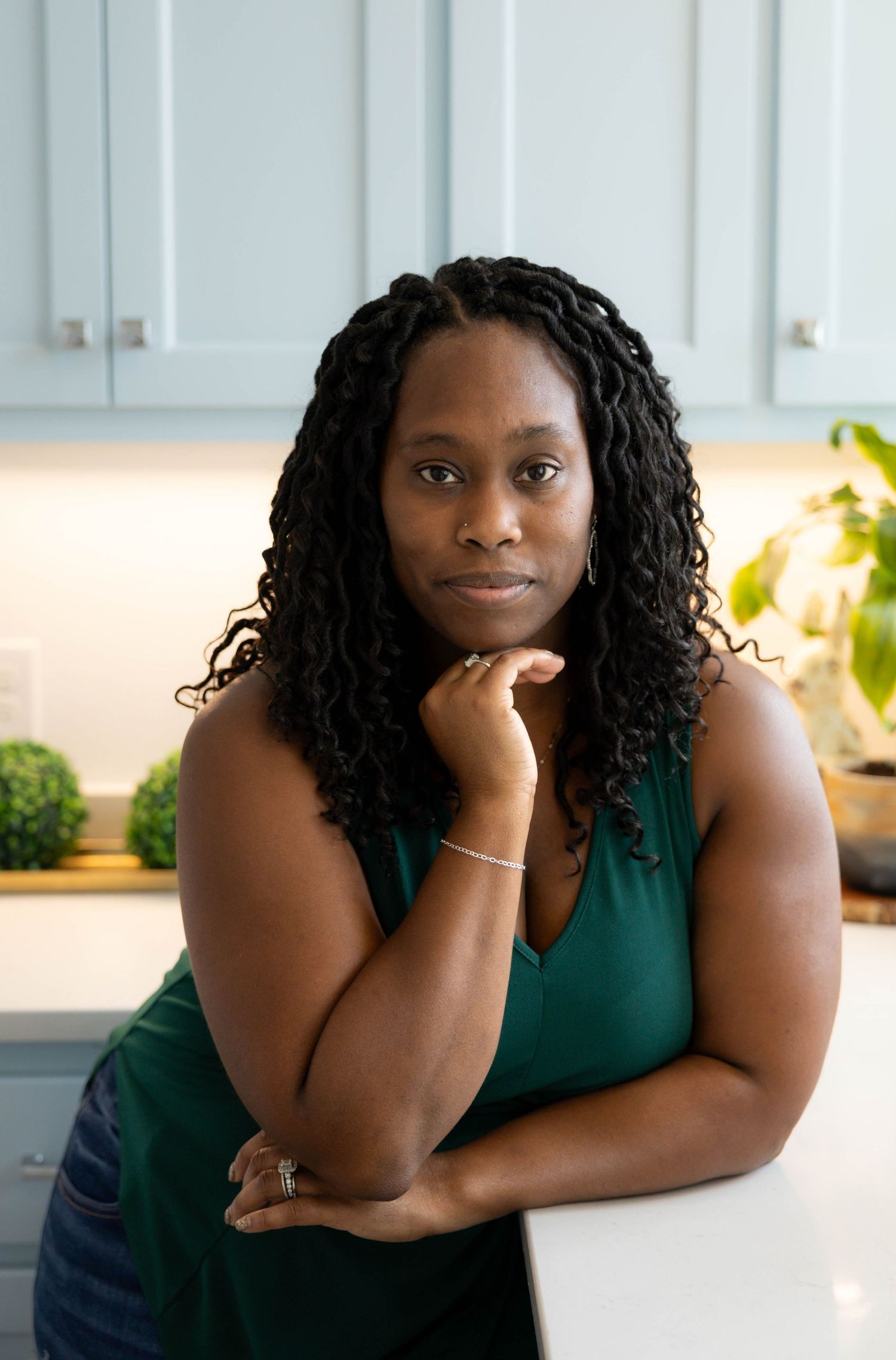 A person in a green sleeveless top rests their chin on their hand while sitting at a table in a bright interior space.