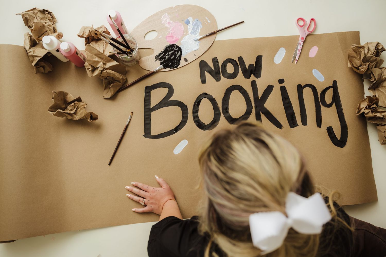 Woman writing Now Booking sign on brown kraft paper with art supplies and crumpled paper surrounding it.