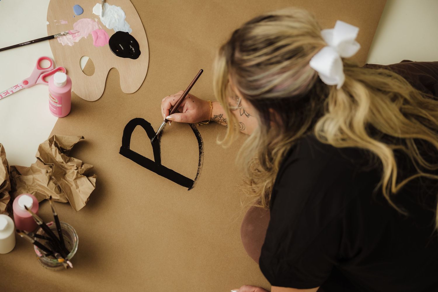 A blonde woman with a white bow writes the letter B in black paint on brown craft paper using a paintbrush.