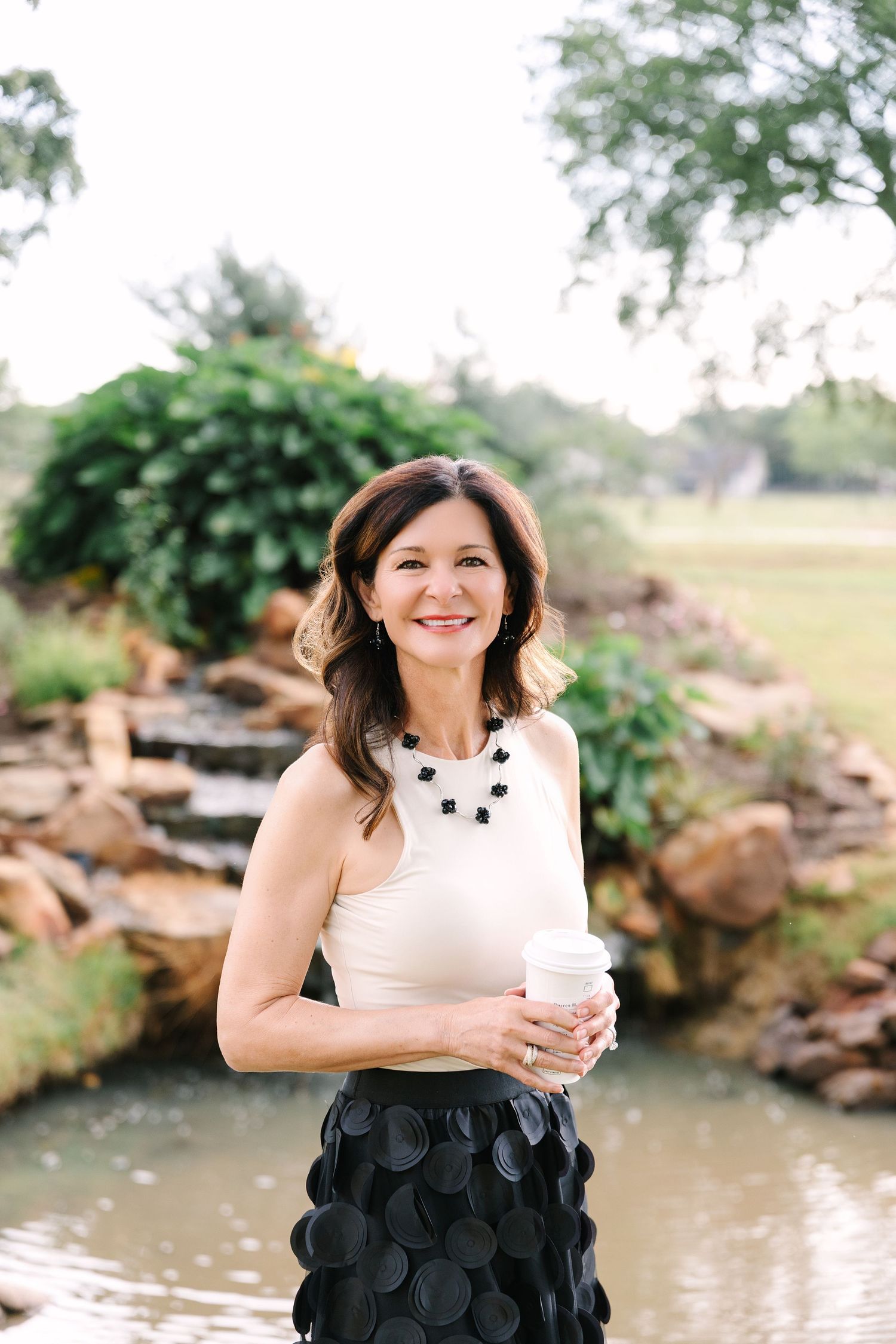 Smiling woman in cream top and black circle skirt holding coffee cup outdoors near a garden pond.