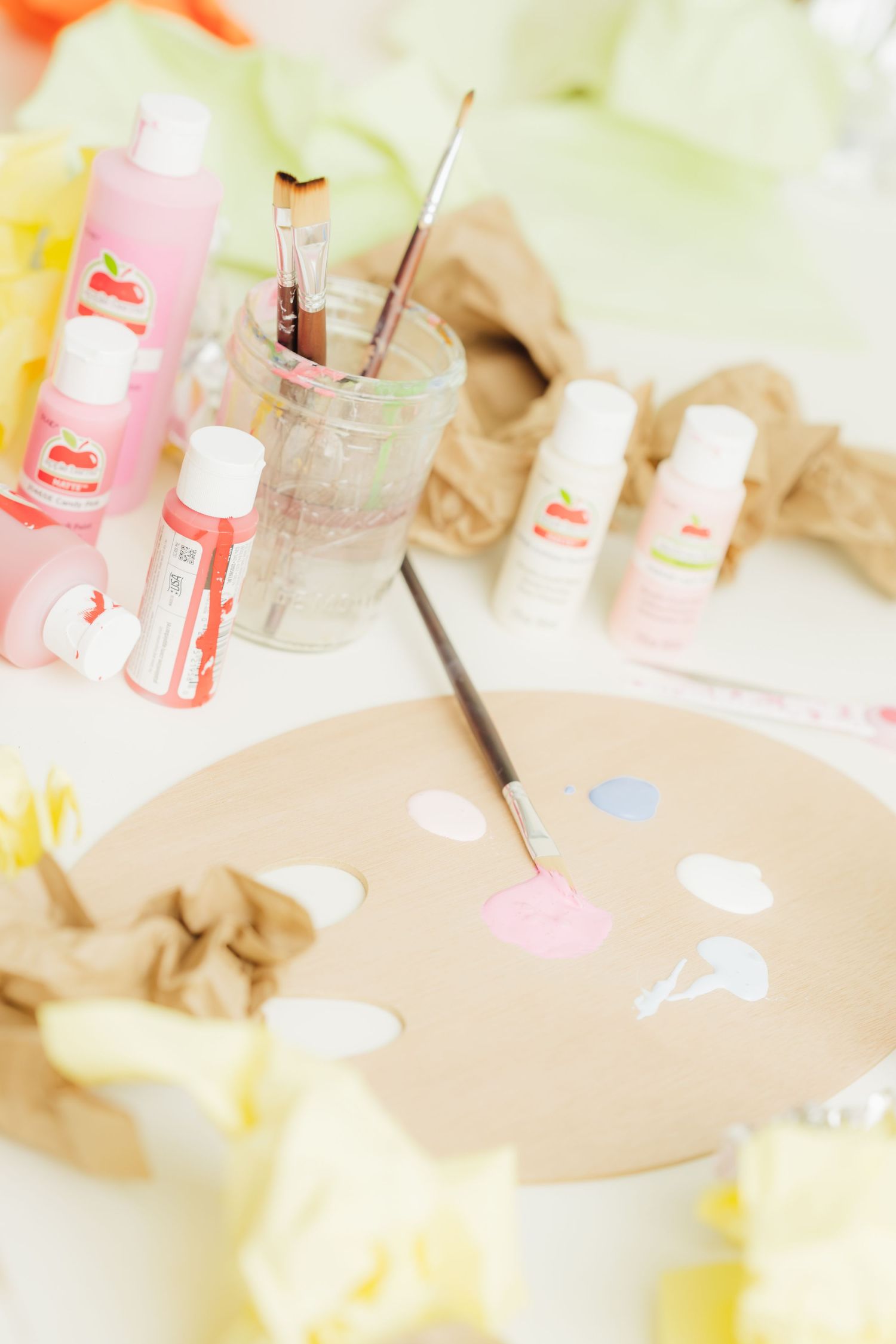 Pastel paint palette with brushes, pink and green acrylic paint bottles, and crinkled paper on a white table.