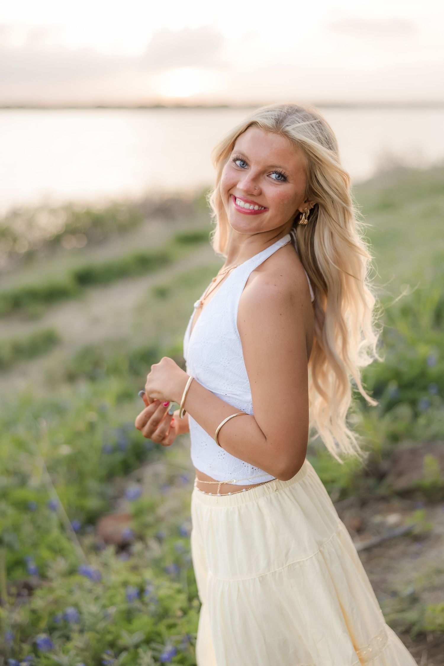 Smiling blonde woman in white top and yellow skirt poses near a lakeside field of wildflowers at sunset.