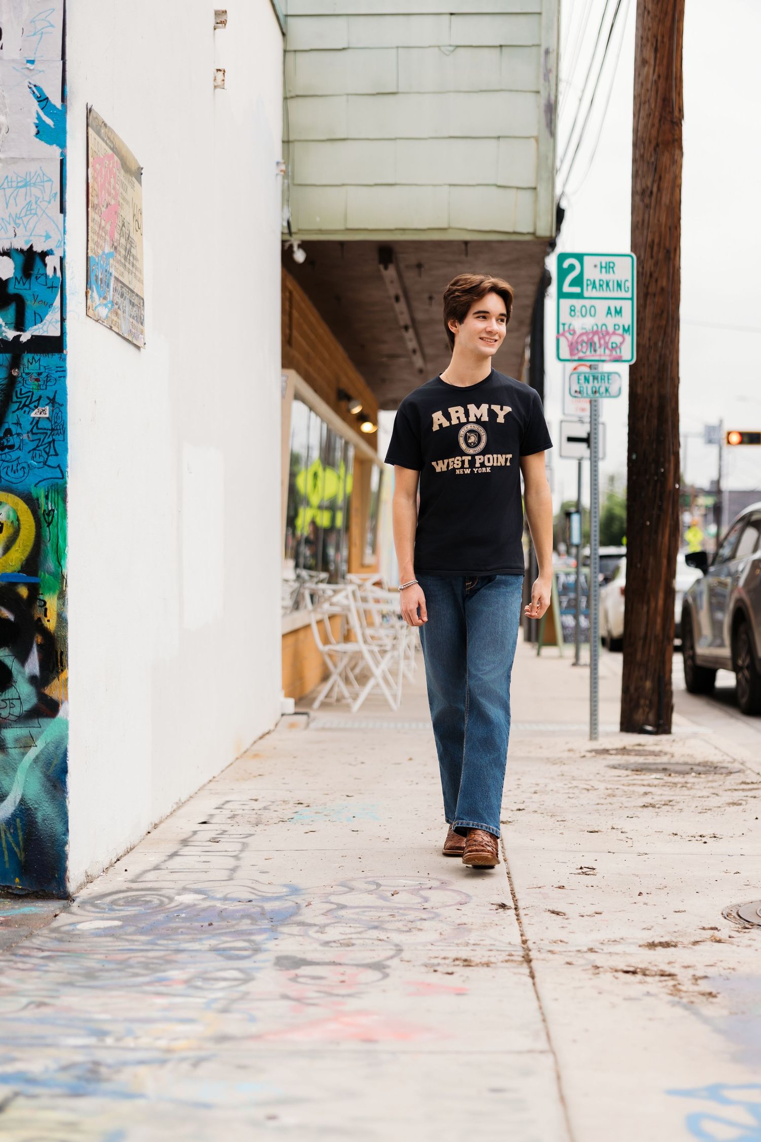 Young man in Army West Point t-shirt and jeans walking on urban sidewalk with graffiti wall.