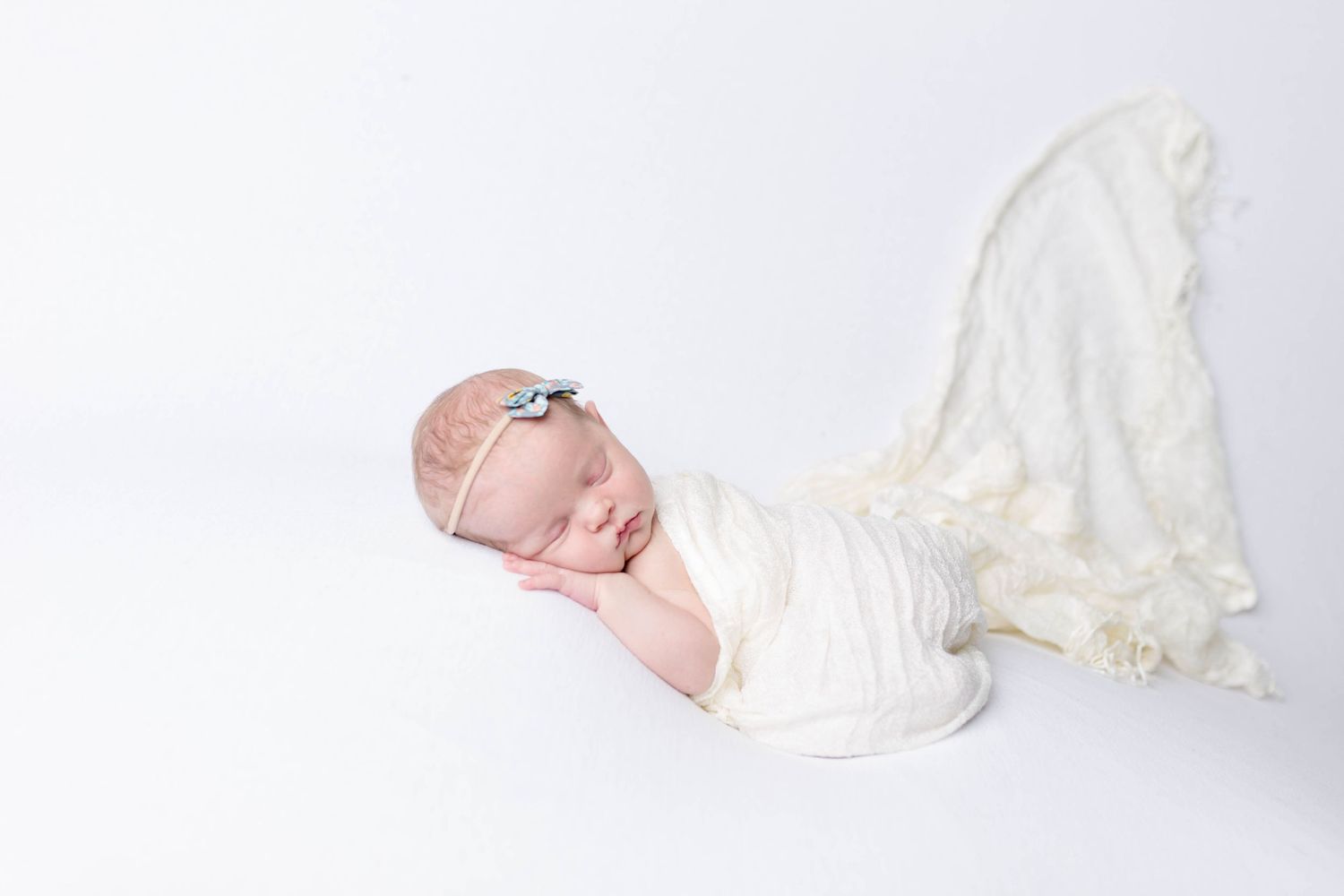 Sleeping newborn baby wrapped in cream colored fabric on white background with delicate headband.