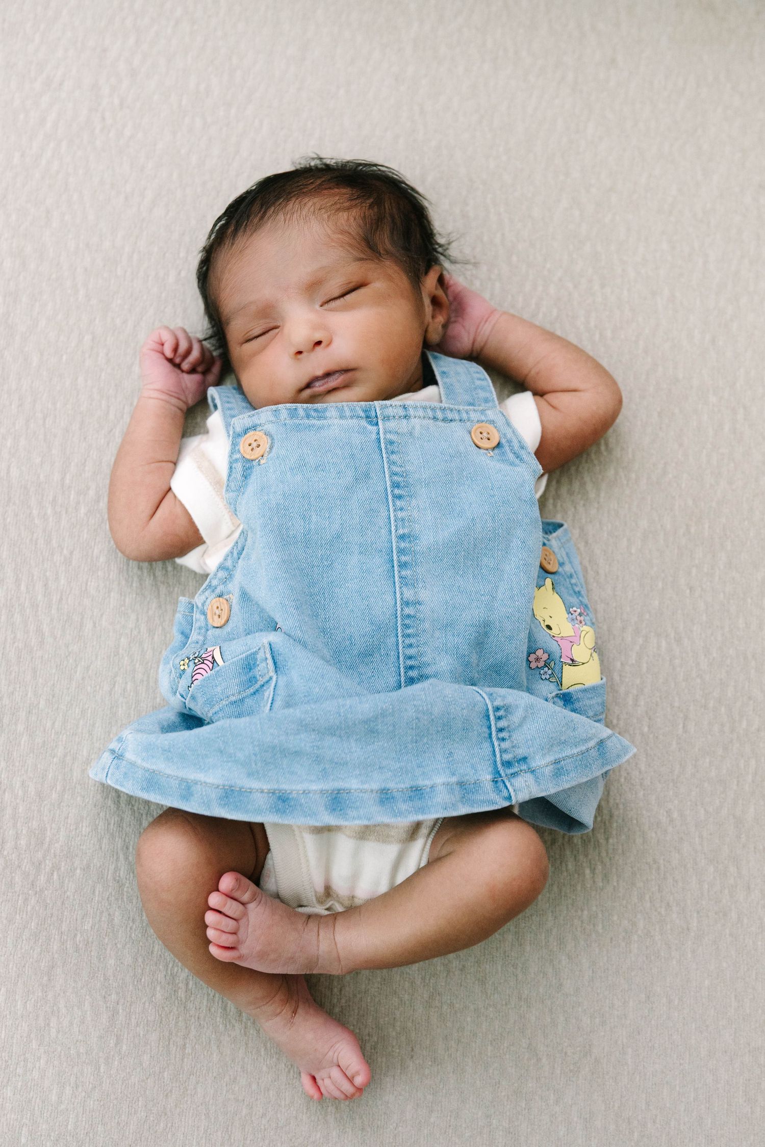 Sleeping newborn baby girl wearing a denim jumper dress lying on a light blanket with hands raised beside her head.