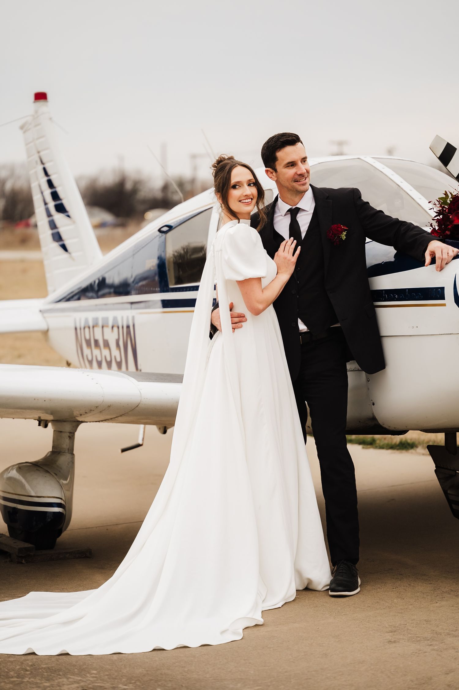 Romantic wedding couple poses next to a small private aircraft on an airfield during their special day.