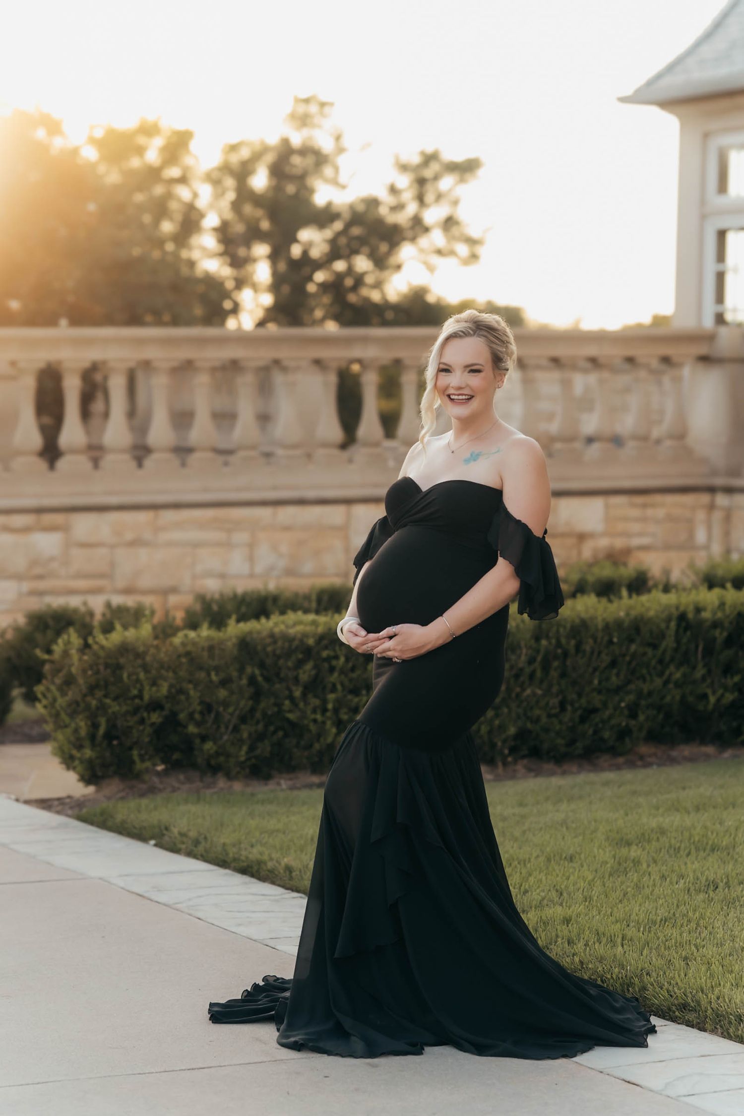 A person in a flowing black dress poses elegantly on stone steps during golden hour at a grand estate.