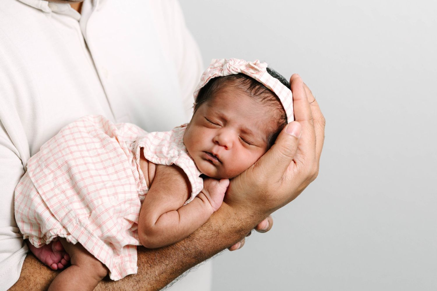 Newborn baby sleeping peacefully in fathers hands, wearing pink outfit and headband.