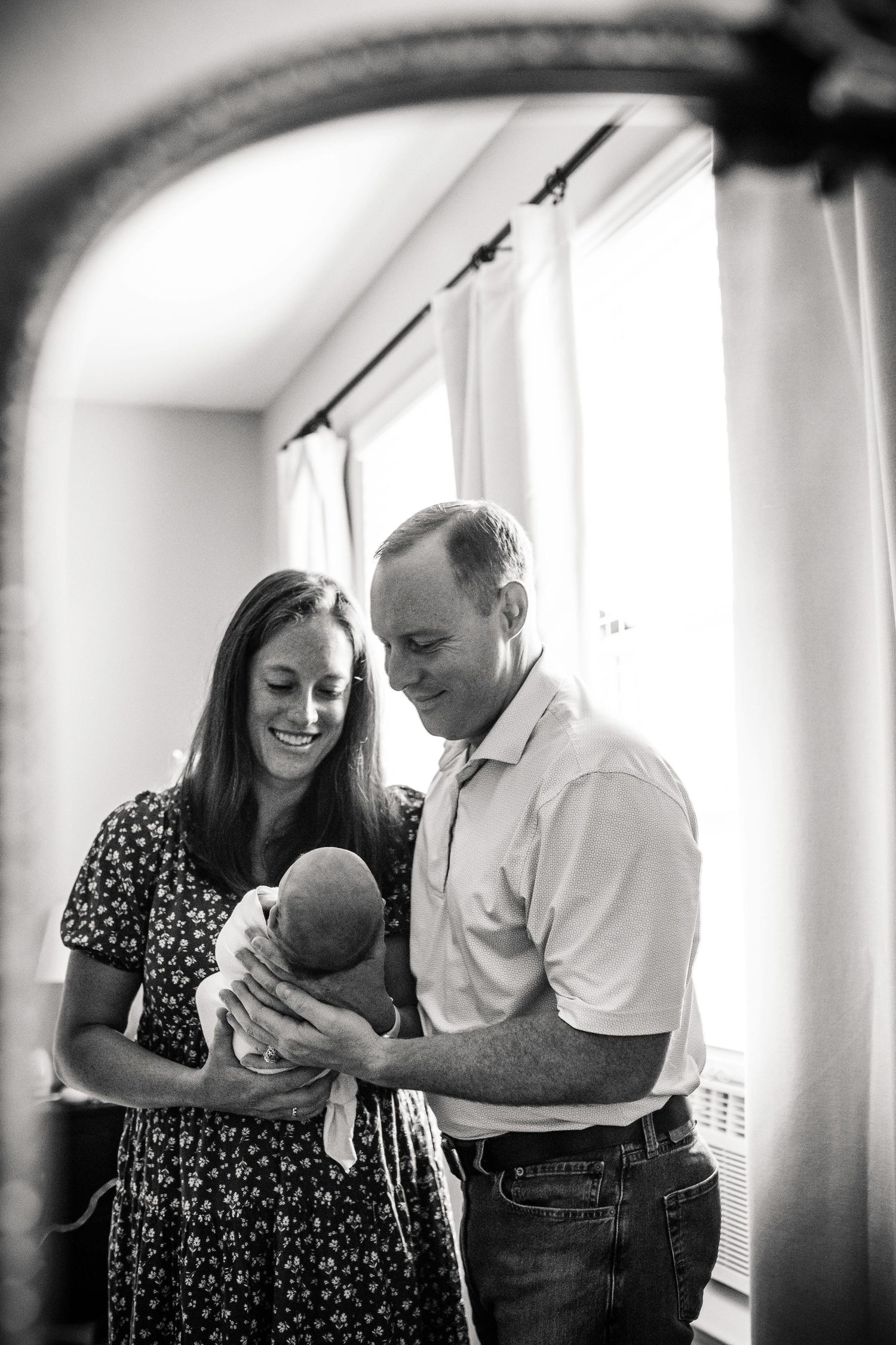 A tender series of black and white photos showing a couple sharing intimate moments with a baby near a bright window.