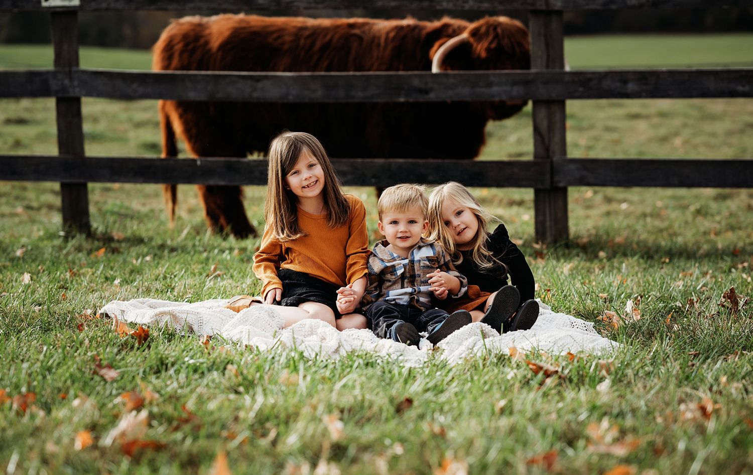 Fall family gathering by a wooden fence with horses grazing in a pastoral setting.