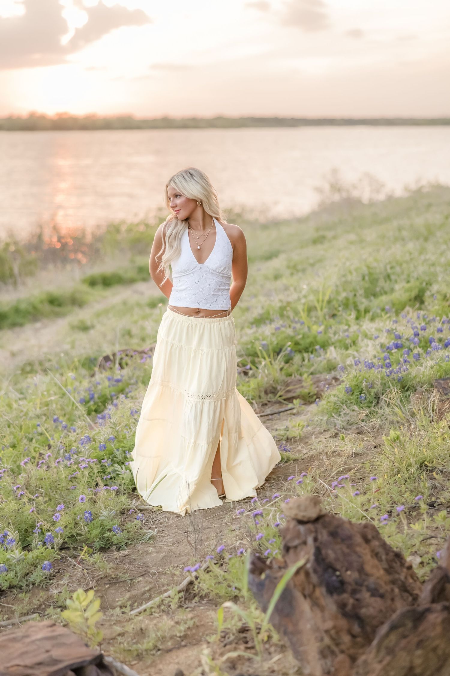 Blonde woman in white crop top and yellow maxi skirt standing near a lake at sunset with wildflowers.