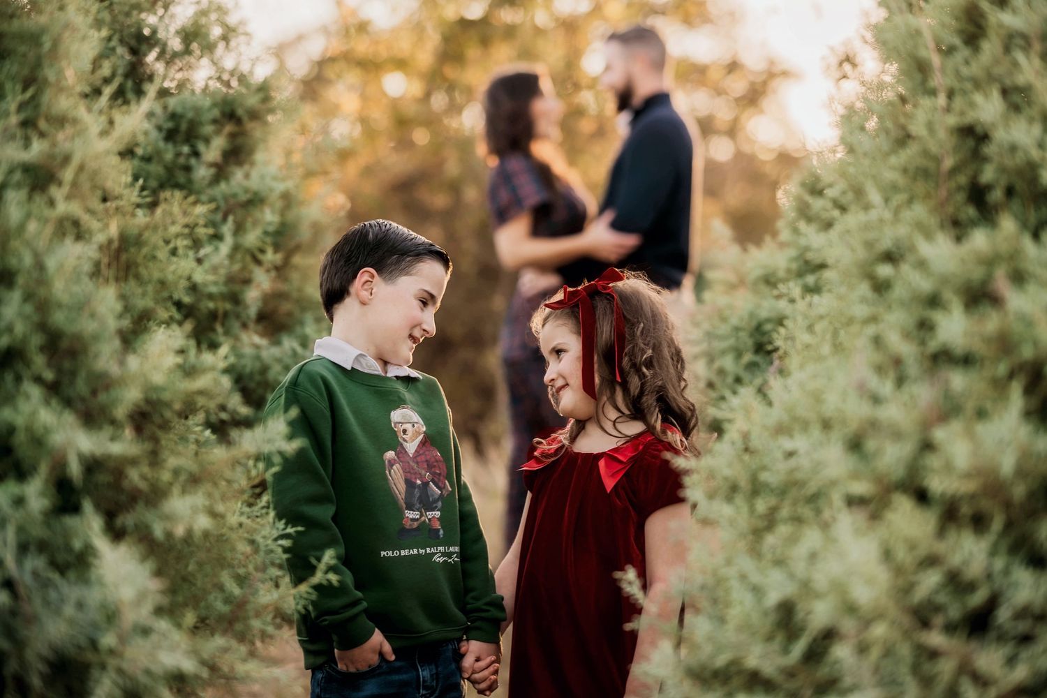 Two children share a sweet moment in a garden while a couple embraces in the blurred background at sunset.