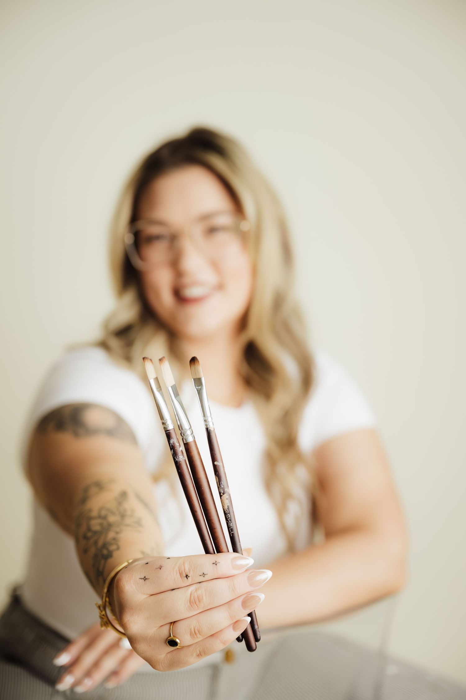 Smiling blonde woman with tattoos holding paint brushes toward camera in white t-shirt against neutral background.