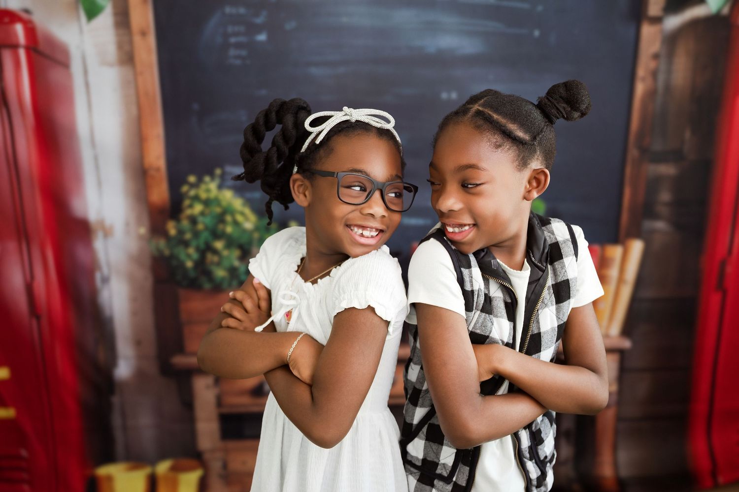 Two young girls smile and pose together in front of a chalkboard, one wearing glasses and a white dress.