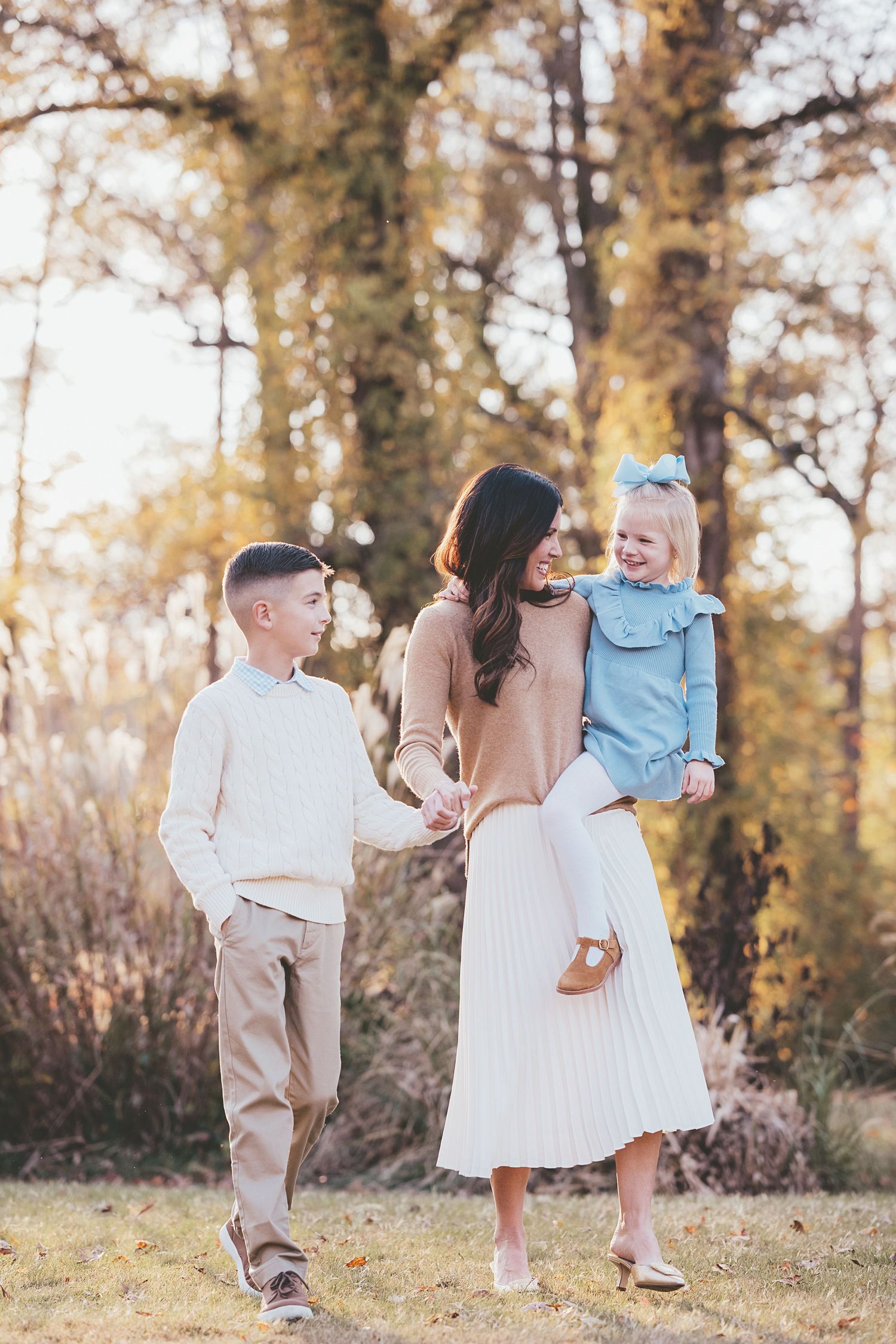 Family wearing coordinated cream and blue outfits walking through autumn woods at sunset.