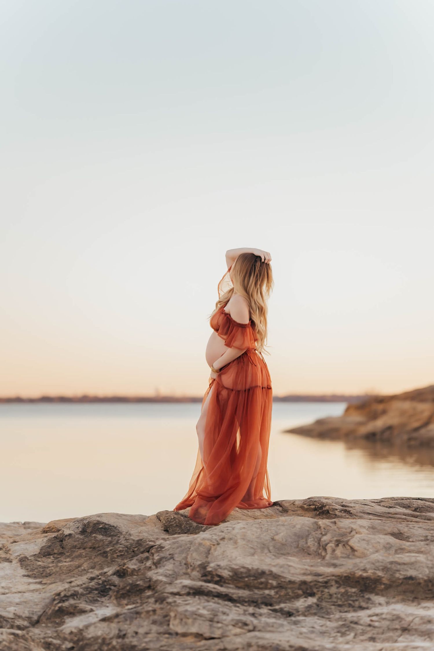 A person in a flowing rust-colored dress sits on coastal rocks at sunset overlooking a serene ocean view.